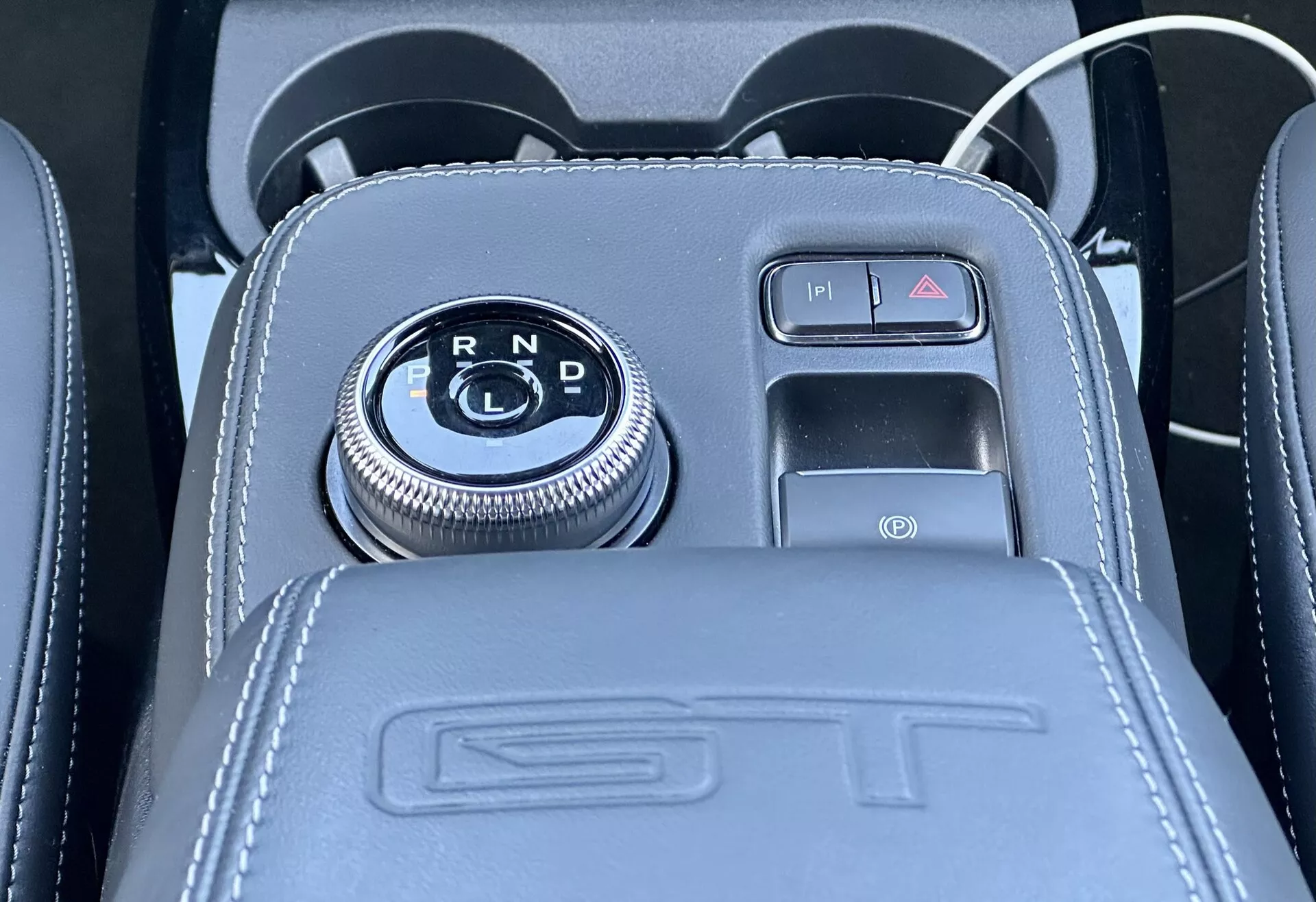 Vue rapprochée de la console centrale de la Ford Mustang Mach-E 2020 présentant un sélecteur de vitesse rotatif sur un panneau noir brillant avec indicateurs de vitesse R, N, D, entouré d'une finition chromée, à côté d'un frein de stationnement électronique à bouton avec détails en cuir, embossage ST, et incluant un port USB et une prise d'accessoires 12V