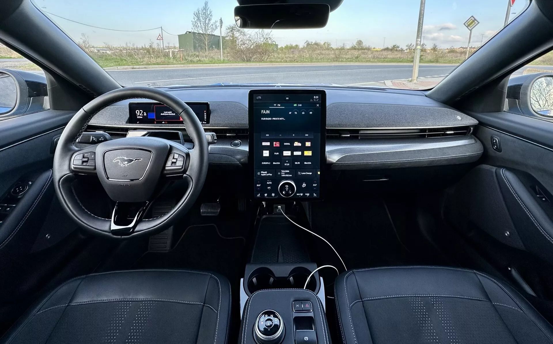 Vue à la première personne de l'intérieur de la Ford Mustang Mach-E 2020, présentant le volant positionné à gauche avec un design à fond plat et emblème, cluster d'instruments numérique, grand système d'infodivertissement tactile vertical sur la console centrale avec boutons de contrôle climatique en dessous, ports USB, et sellerie de luxe en tons foncés avec coutures détaillées, suggérant des caractéristiques de sécurité et d'assistance avancées, avec un arrière-plan extérieur rural visible à travers le pare-brise.