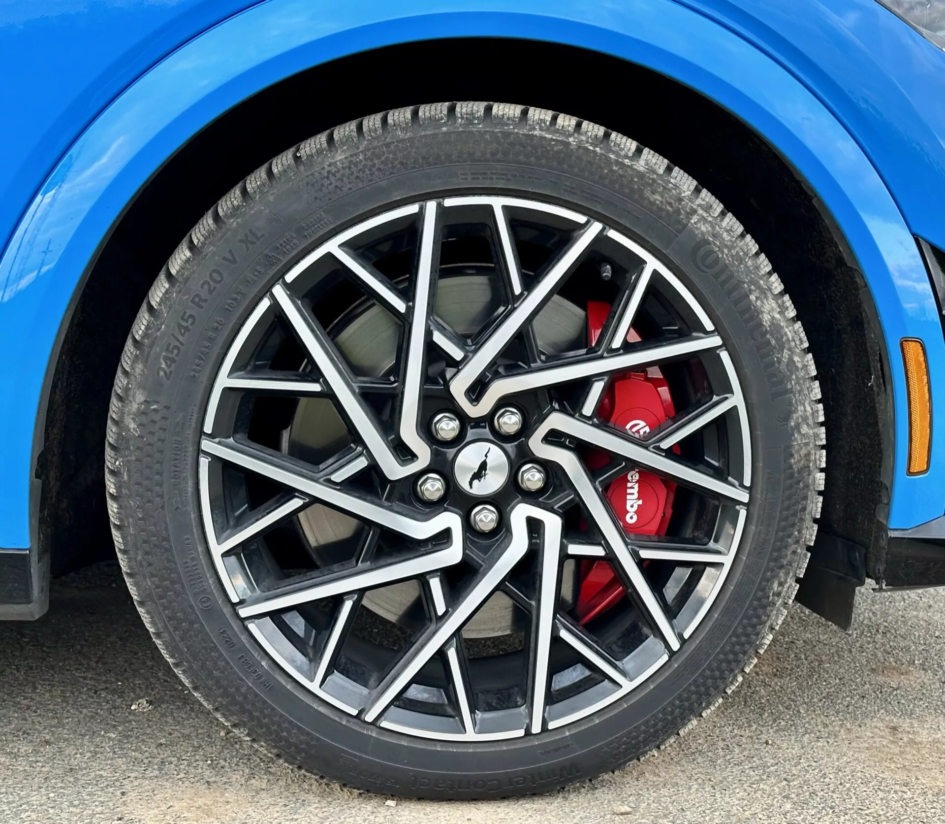 Gros plan sur la roue avant droite du Ford Mustang Mach-E 2020 avec jante en alliage à multiples rayons et système de freinage haute performance avec étrier rouge, présentant les détails du pneu toutes saisons et un segment de l'aile bleu vif, pris sous un angle bas.