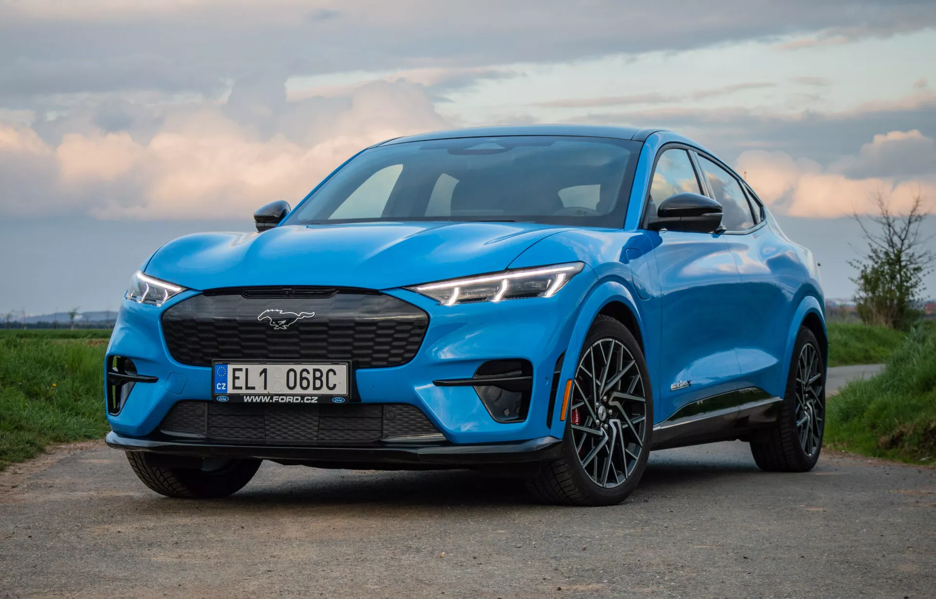 Ford Mustang Mach-E bleu 2020 photographié sous un angle bas et légèrement décentré, montrant la grille, les phares, le capot, et le profil du SUV électrique incluant la roue avant et l'aile, soulignant son design performant et aérodynamique à l'aube ou au crépuscule.