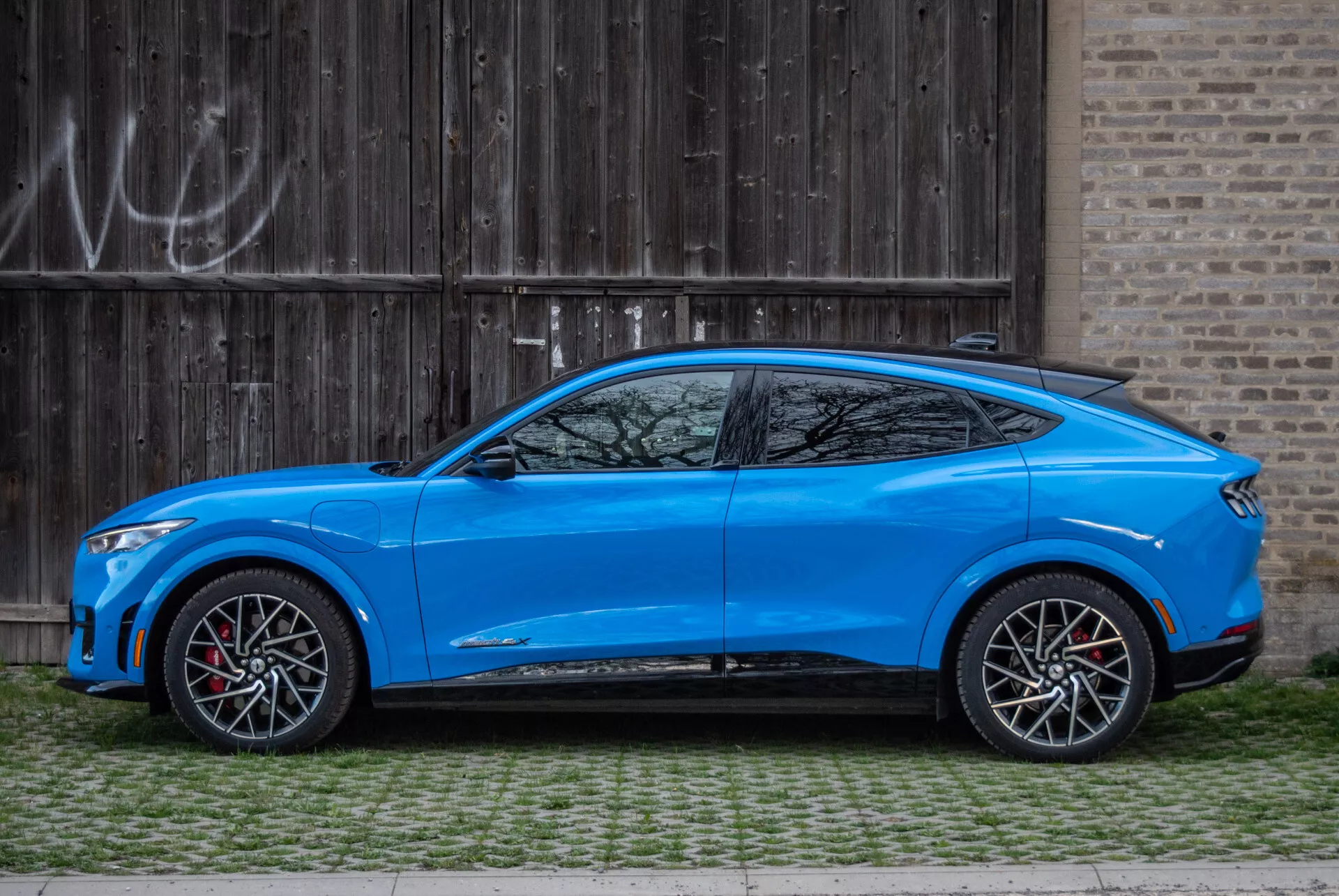 SUV crossover bleu Ford Mustang Mach-E 2020 garé parallèlement à une porte de garage en bois rustique avec graffitis, vue de profil mettant en évidence la conception aérodynamique, ligne de toit inclinée de style coupé, modèle quatre portes avec poignées intégrées, rétroviseurs latéraux stylés aérodynamiquement avec clignotants, grandes roues en alliage bicolores avec étriers de frein rouges, garde au sol modérée, carrosserie profilée avec lignes sculptées et passages de roue prononcés, ligne de vitres effilées avec petite fenêtre de quart après le pilier C et jupes latérales sportives