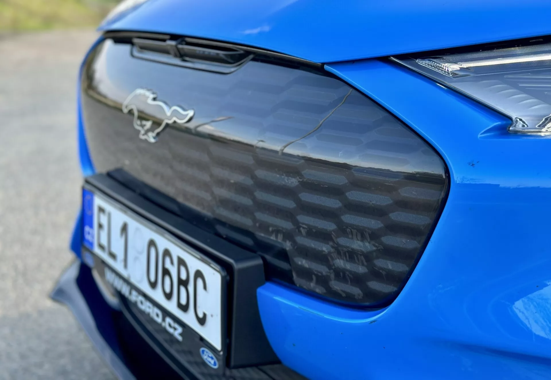 Gros plan sur la section arrière d'une Ford Mustang Mach-E 2020 bleue, mettant en avant l'emblème de cheval chromé, l'aileron arrière en fibre de carbone, le logement intégré pour la plaque d'immatriculation et un aperçu partiel des feux arrière LED, soulignant les caractéristiques aérodynamiques et de performance de la voiture.