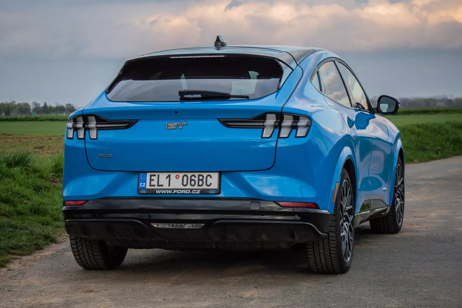 Ford Mustang-Mach-E 2020 photographiée sous un angle arrière et latéral mettant en évidence son design distinctif, incluant des feux arrière LED horizontaux, l'emblème du fabricant, un aileron arrière intégré, un pare-chocs arrière équipé de capteurs sans échappement visible suggérant un statut de véhicule électrique, une plaque d'immatriculation européenne, des jantes en alliage à multiples branches, une peinture bleue vibrante et une carrosserie profilée aérodynamique, sur un fond de campagne.