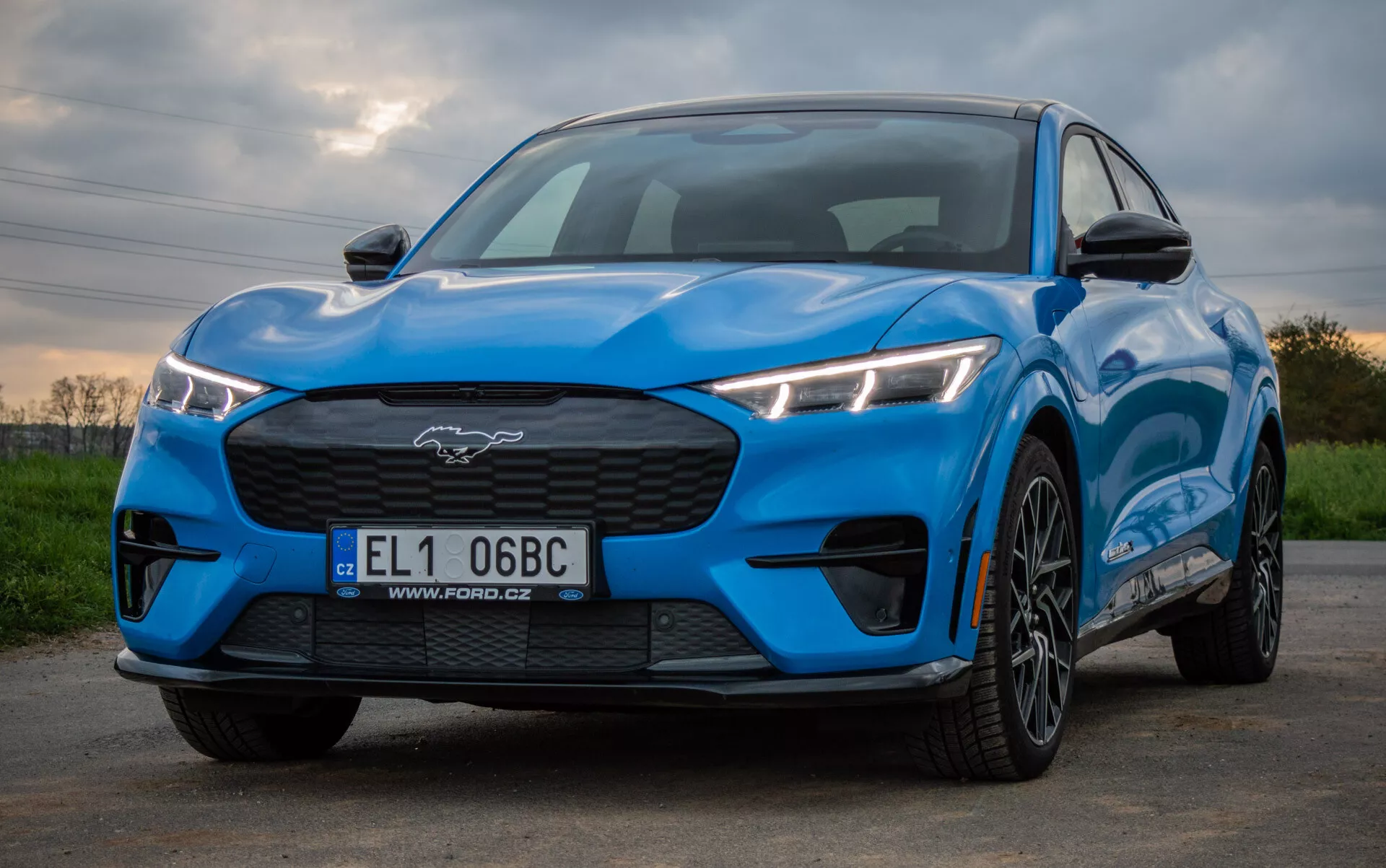 Ford Mustang Mach-E 2020 en bleu vif présentant un design sportif et aérodynamique depuis une perspective avant-trois-quarts basse, mettant en évidence des phares LED angulaires, une grille fermée, un capot profilé et des roues en alliage à multiples branches sur un fond naturel.