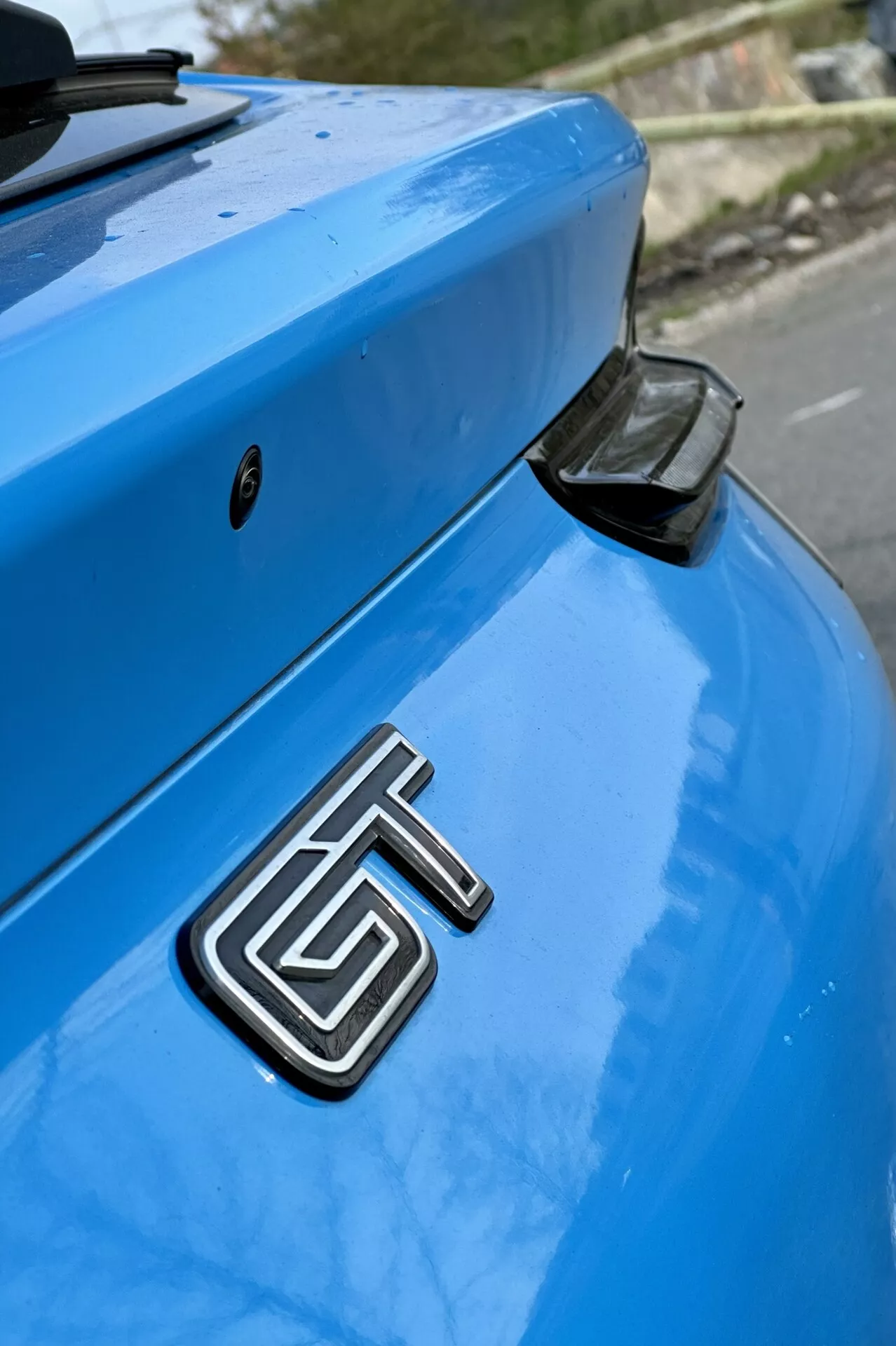 Gros plan sur la section arrière d'une Ford Mustang Mach-E bleue 2020 mettant en évidence le badge métallique GT, le coffre incurvé et l'aileron arrière aérodynamique avec un capuchon d'extrémité noir, le tout sur un arrière-plan légèrement flou.