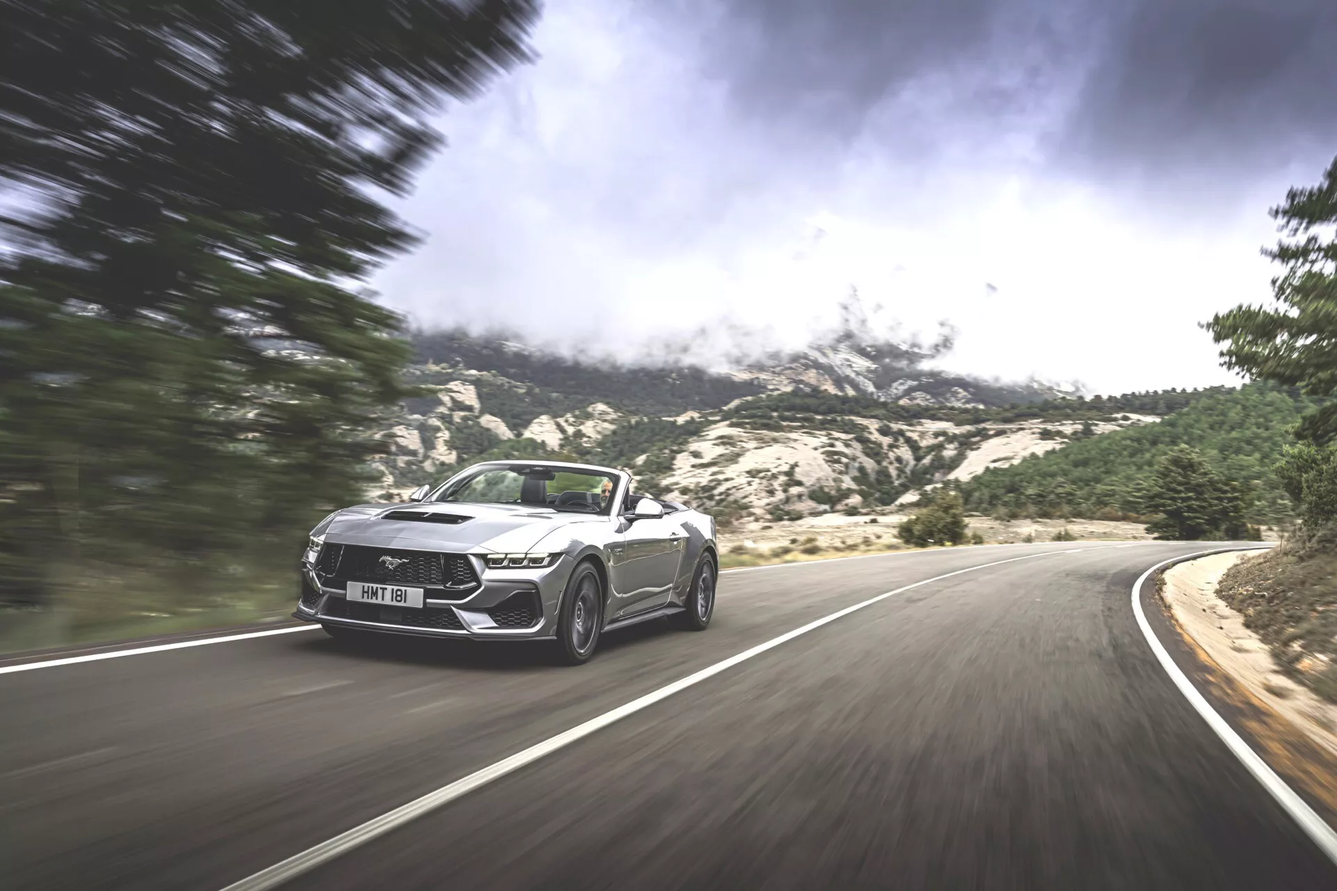 Cliché dynamique d'une Ford Mustang Convertible 2024 en mouvement sur une route sinueuse en montagne, présentant un design sportif et des caractéristiques de luxe sur un fond dramatique.