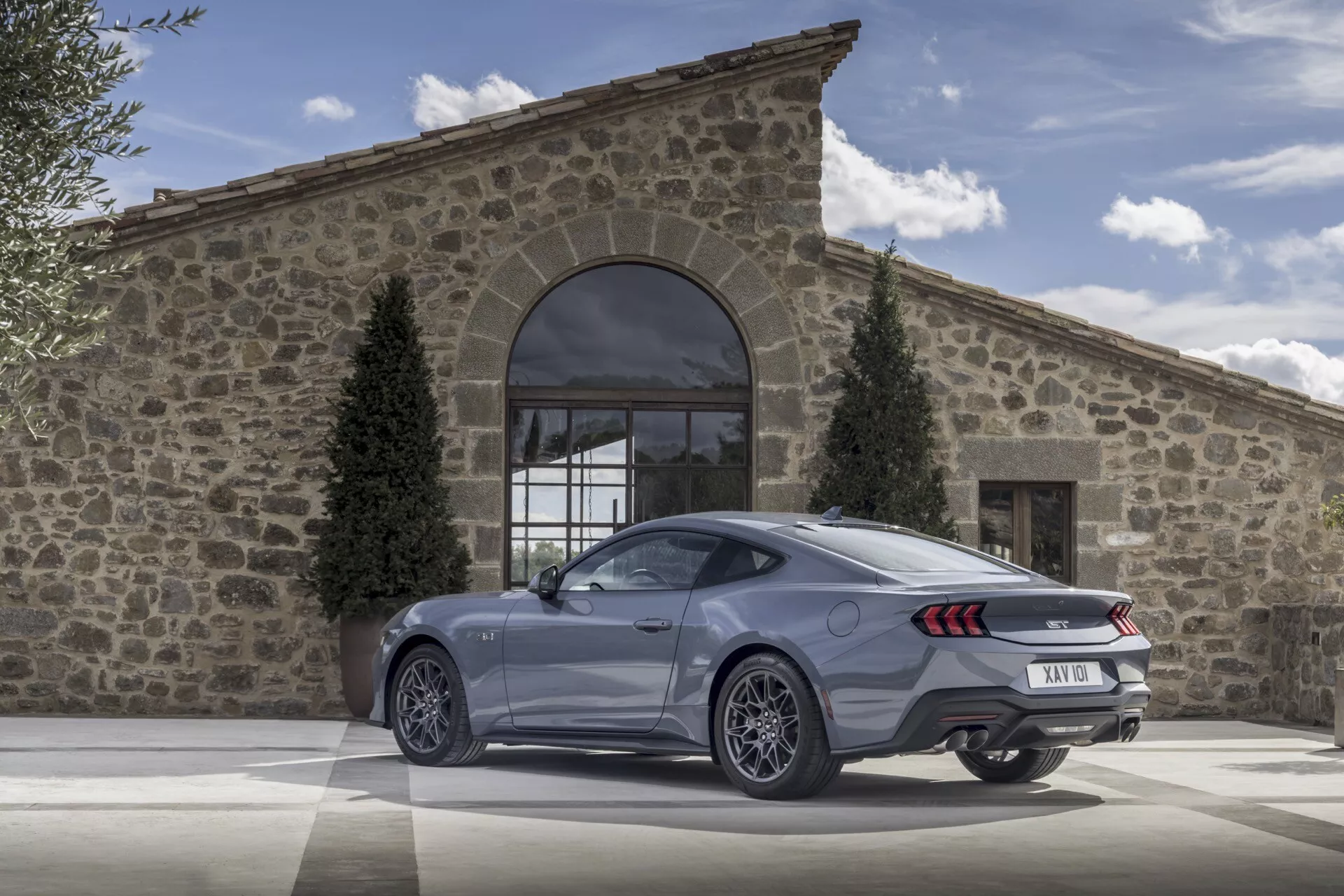Ford Mustang coupé 2024 présentant une vue arrière trois quarts avec feux arrière LED, double échappement et grandes roues en alliage sur une allée pavée devant un bâtiment rustique par une journée claire, mettant en évidence ses caractéristiques de luxe et de performance.
