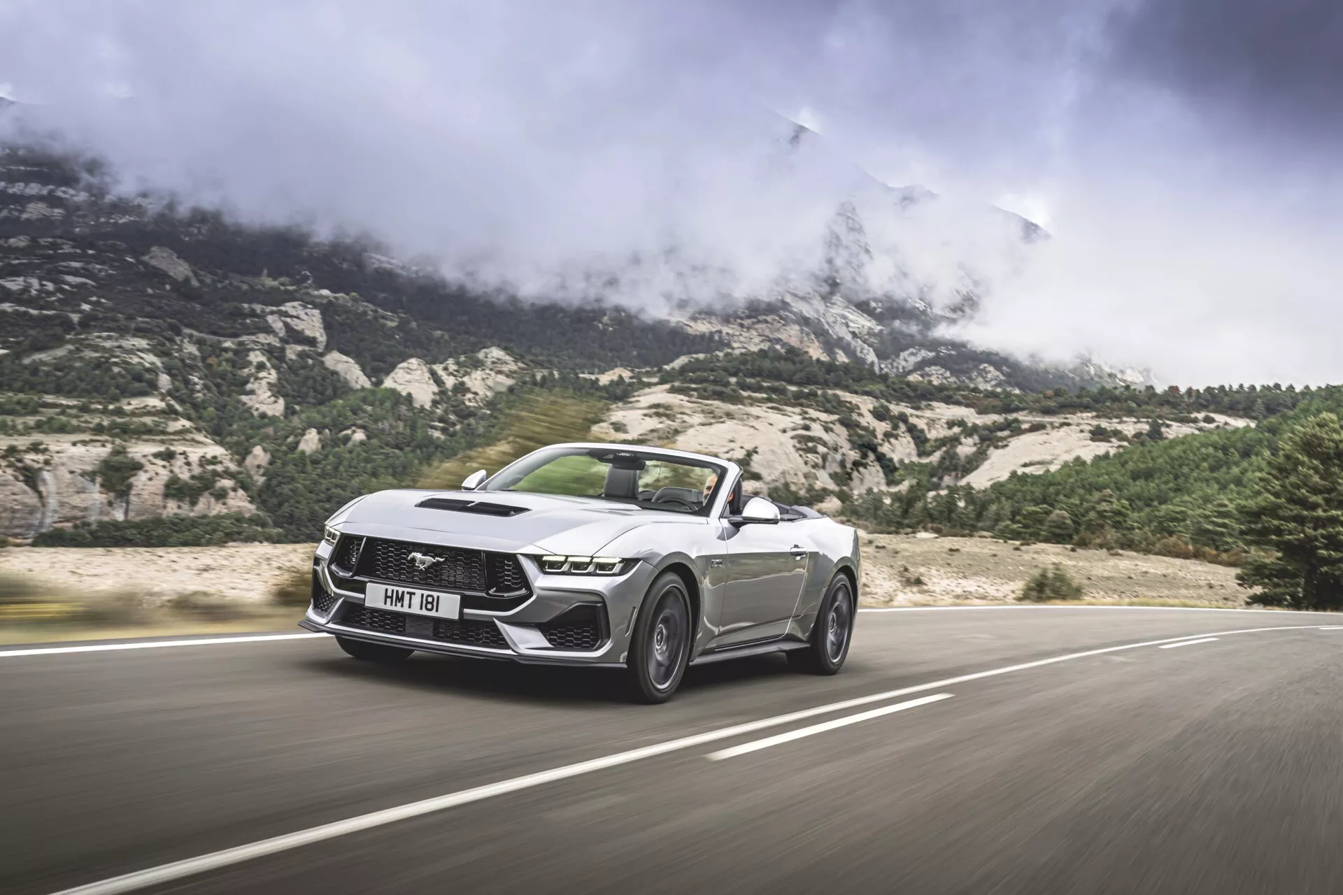 Ford Mustang Cabriolet 2024 en mouvement, montrant ses caractéristiques de design aiguisées et angulaires, y compris une grille proéminente avec un motif distinctif, des phares LED et un capot sculpté, vu sous un angle avant bas de trois quarts avec un fond montagneux