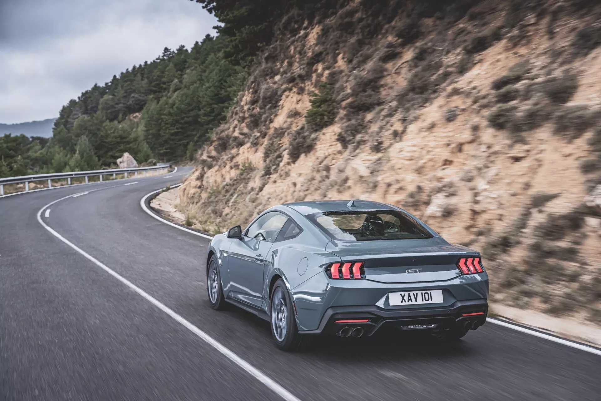Coupé argenté Ford Mustang 2024 sur route sinueuse : Vue arrière 3/4 mettant en lumière les feux arrière LED, double échappement et caractéristiques aérodynamiques sur un fond pittoresque