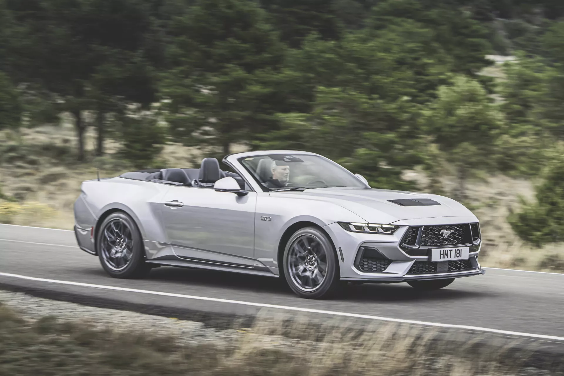 Ford Mustang Convertible 2024 en argent, présentant son style agressif et ses fonctionnalités avancées en mouvement sur une route, capturé depuis une perspective de trois quarts du côté conducteur