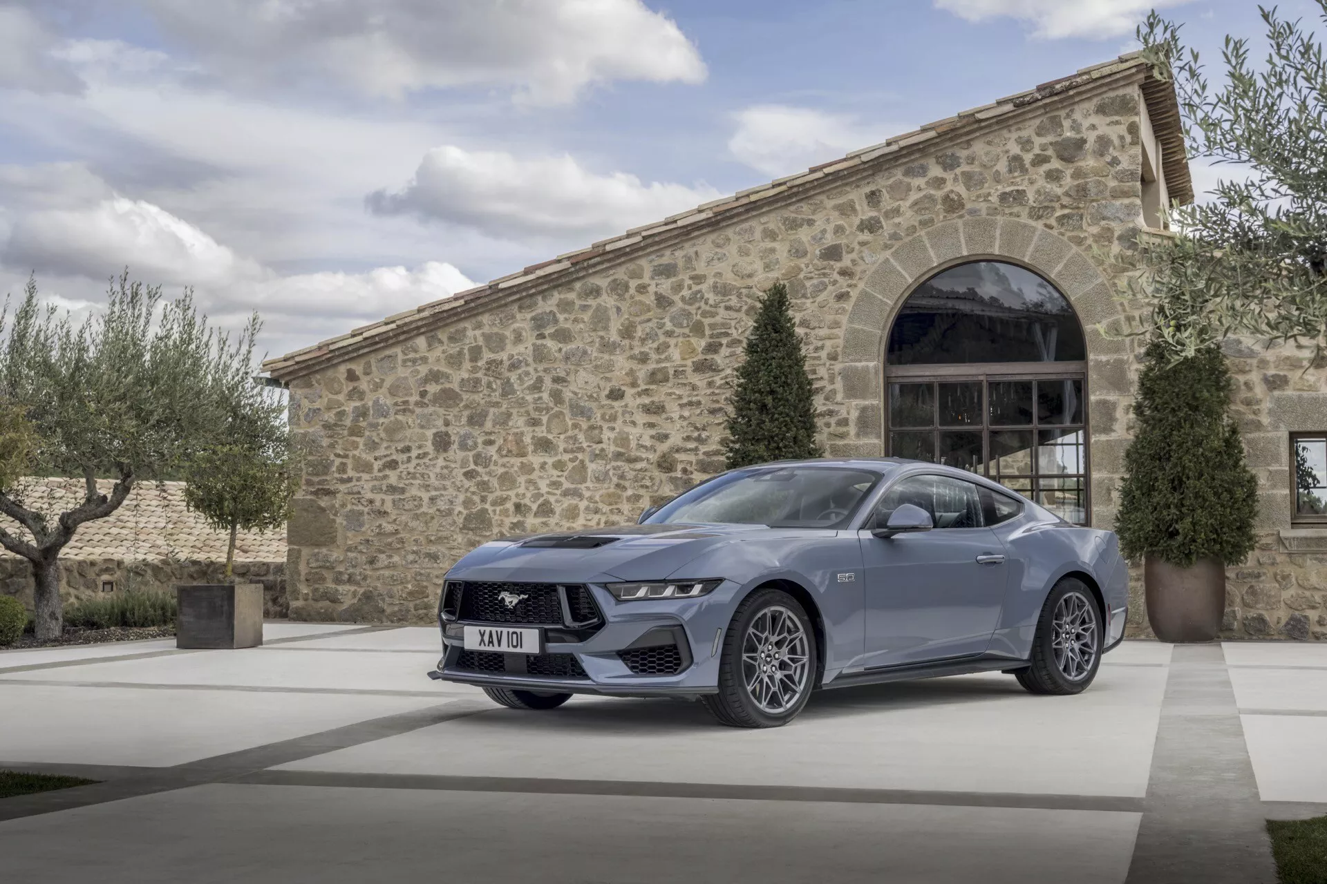 Ford Mustang 2024 à la finition métallique élégante stationnée devant un bâtiment haut de gamme à façade en pierre, affichant son design sportif et élégant avec une vue détaillée de ses phares LED angulaires, de sa grille avant distinctive et de ses roues en alliage à multiples branches