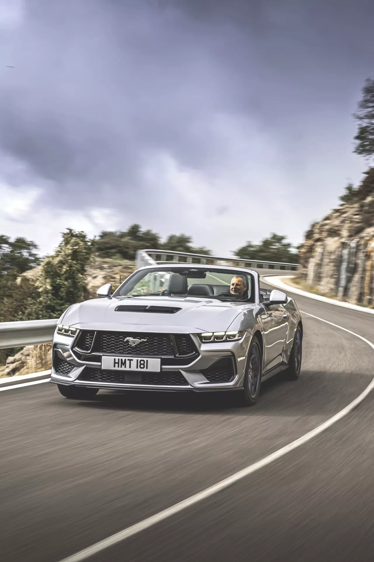Ford Mustang convertible 2024 de 2e génération en conduite dynamique sur route sinueuse présentant des phares LED, une grille emblématique, un toit convertible baissé avec intérieur et conducteur visibles, conçue pour la performance et le luxe