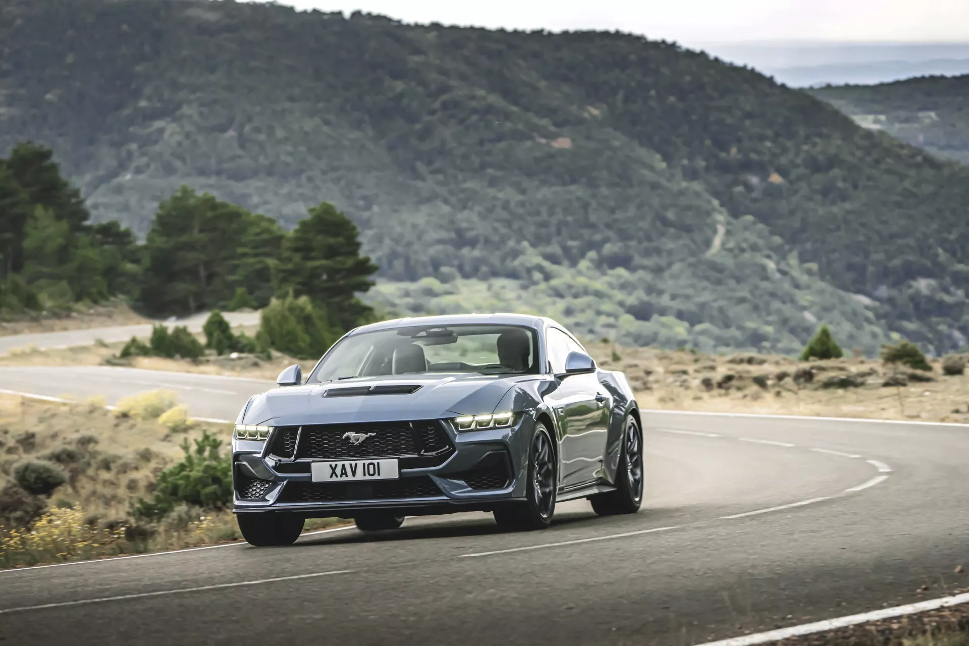 Ford Mustang de 2ème génération 2024 dans une scène de conduite dynamique sur une route montagneuse sinueuse, présentant une posture agressive, des phares LED modernes, une grille complexe, des lignes musculaires sur le capot et des roues en alliage sportives
