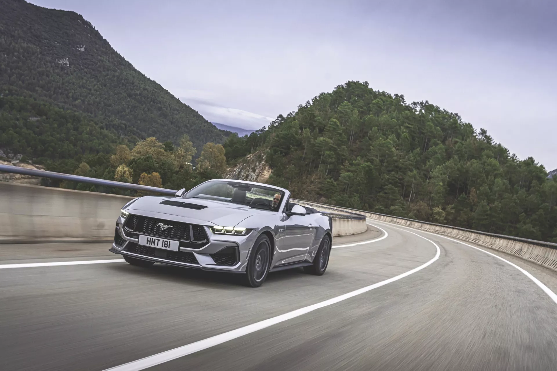 Ford Mustang 2024 en mouvement sur une route de montagne, mettant en avant son design de voiture musclée moderne avec un emblème emblématique de cheval au galop, une façade avant agressive avec des phares élégants, un pare-choc sportif, des lignes sculptées sur le capot, des roues en alliage distinctives et un toit convertible en finition métallique.