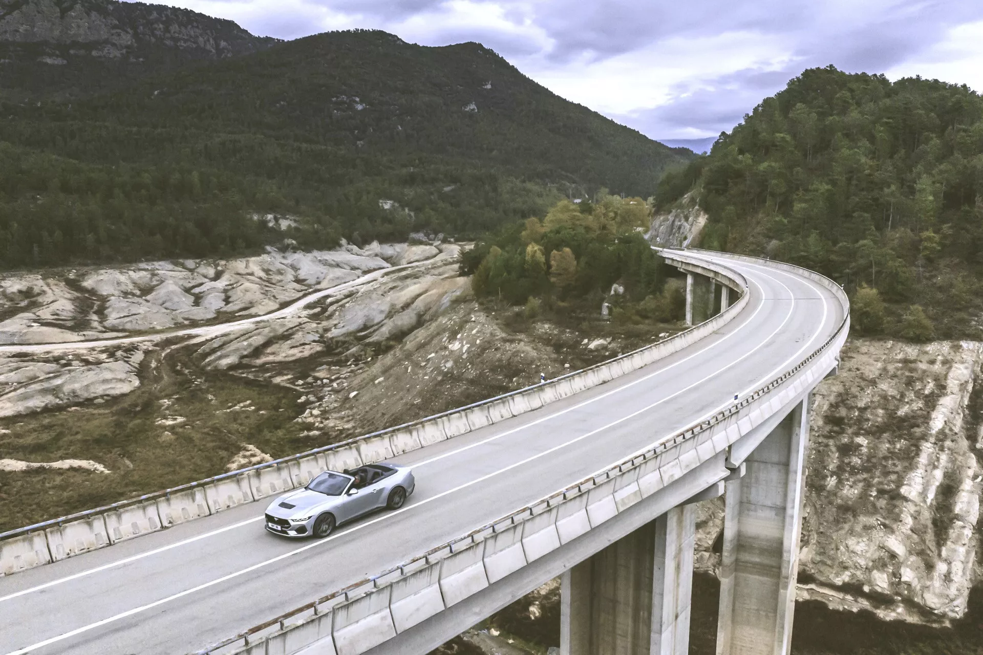 Ford Mustang 2024 voyageant sur un pont dans un paysage vallonné, capturé d'un angle élevé montrant le côté avant gauche et la vue de dessus avec un focus sur la peinture métallique et les caractéristiques de conception comme la grille avant et les roues en alliage sous une lumière naturelle