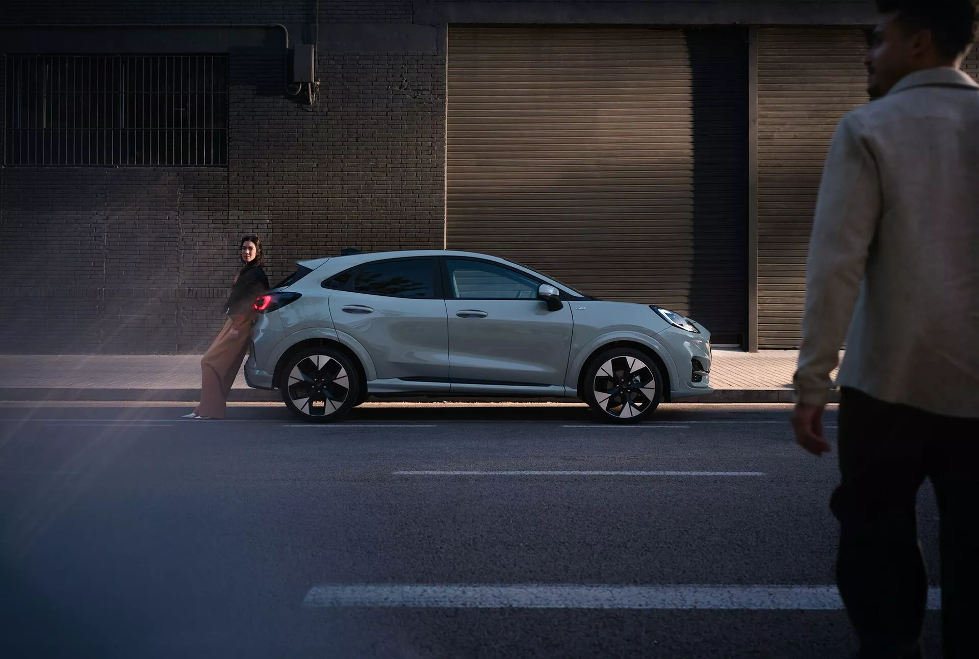 Ford Puma 2024 (II-facelift) hatchback en argent métallisé avec un design sportif, capturée depuis une perspective de niveau de rue montrant son profil élégant et des caractéristiques modernes telles que des détails noirs et des roues à plusieurs branches sur un fond urbain