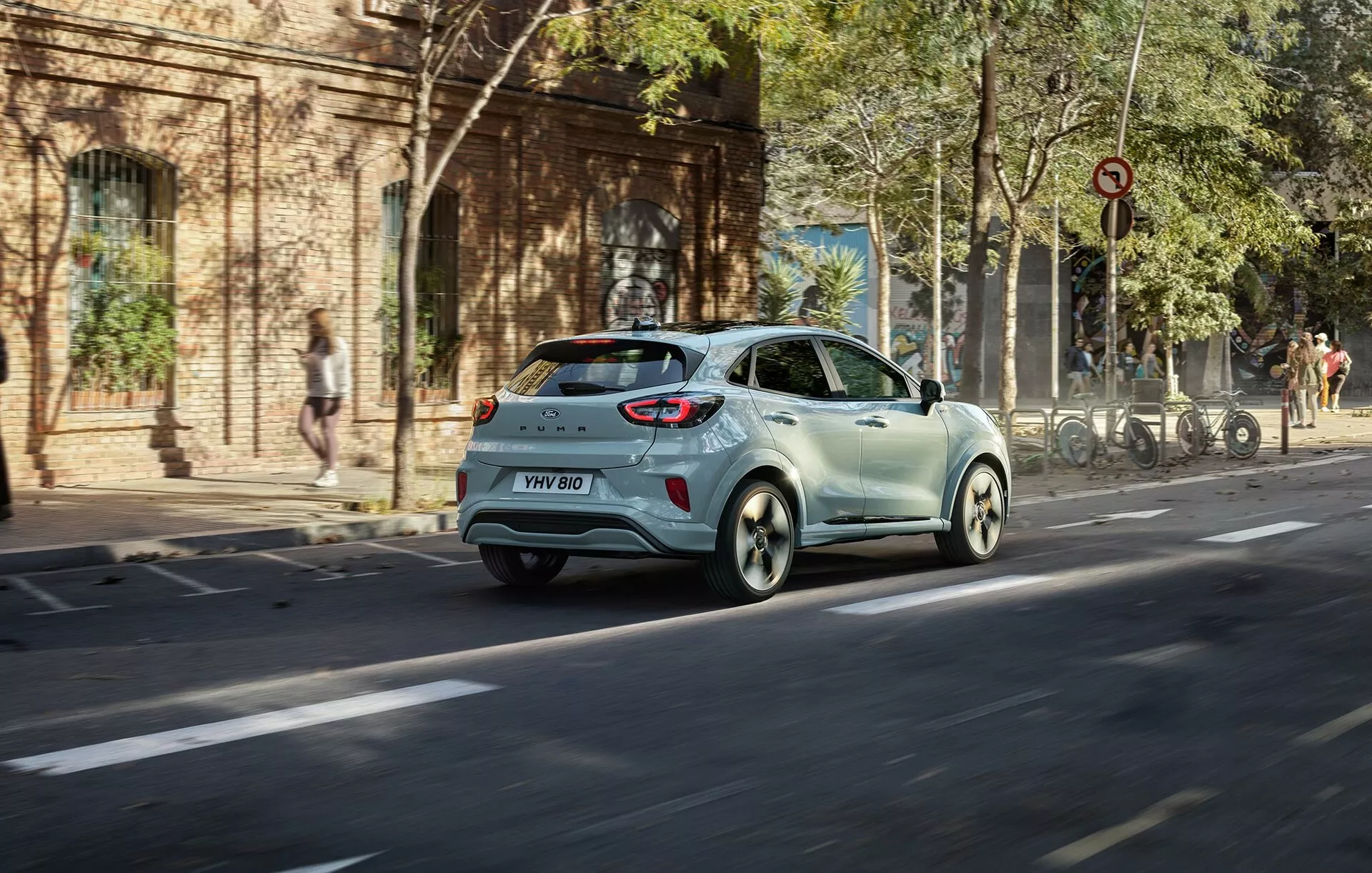 Ford Puma 2024 (II-facelift) hatchback de couleur claire en mouvement dans une rue de la ville depuis un angle arrière de trois quarts montrant les feux arrière, la fenêtre arrière, les portes latérales et les roues en alliage