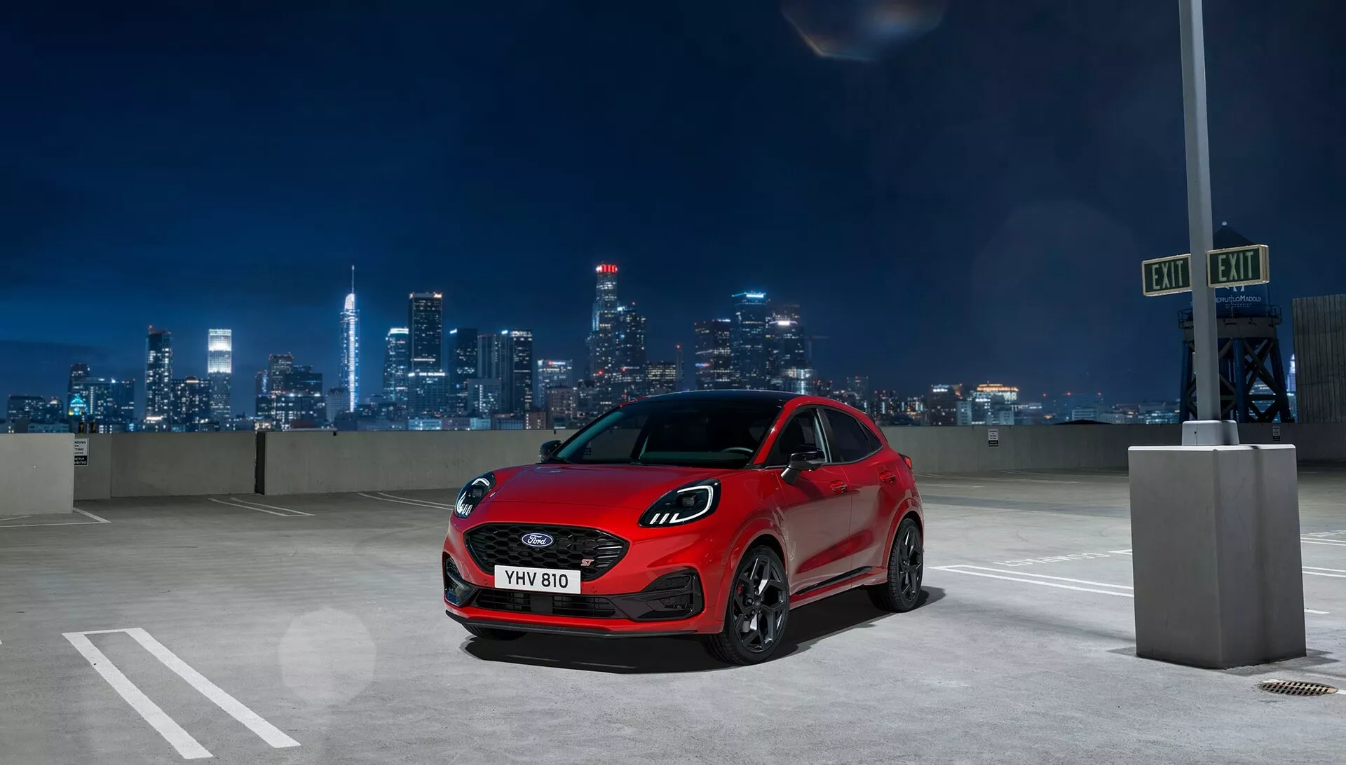 Hatchback rouge Ford Puma 2024 (II facelift) garée sur un parking en plein air la nuit avec un horizon de ville illuminé en arrière-plan, montrant des phares LED agressifs, une grande calandre et un profil sportif depuis une vue avant basse à trois quarts