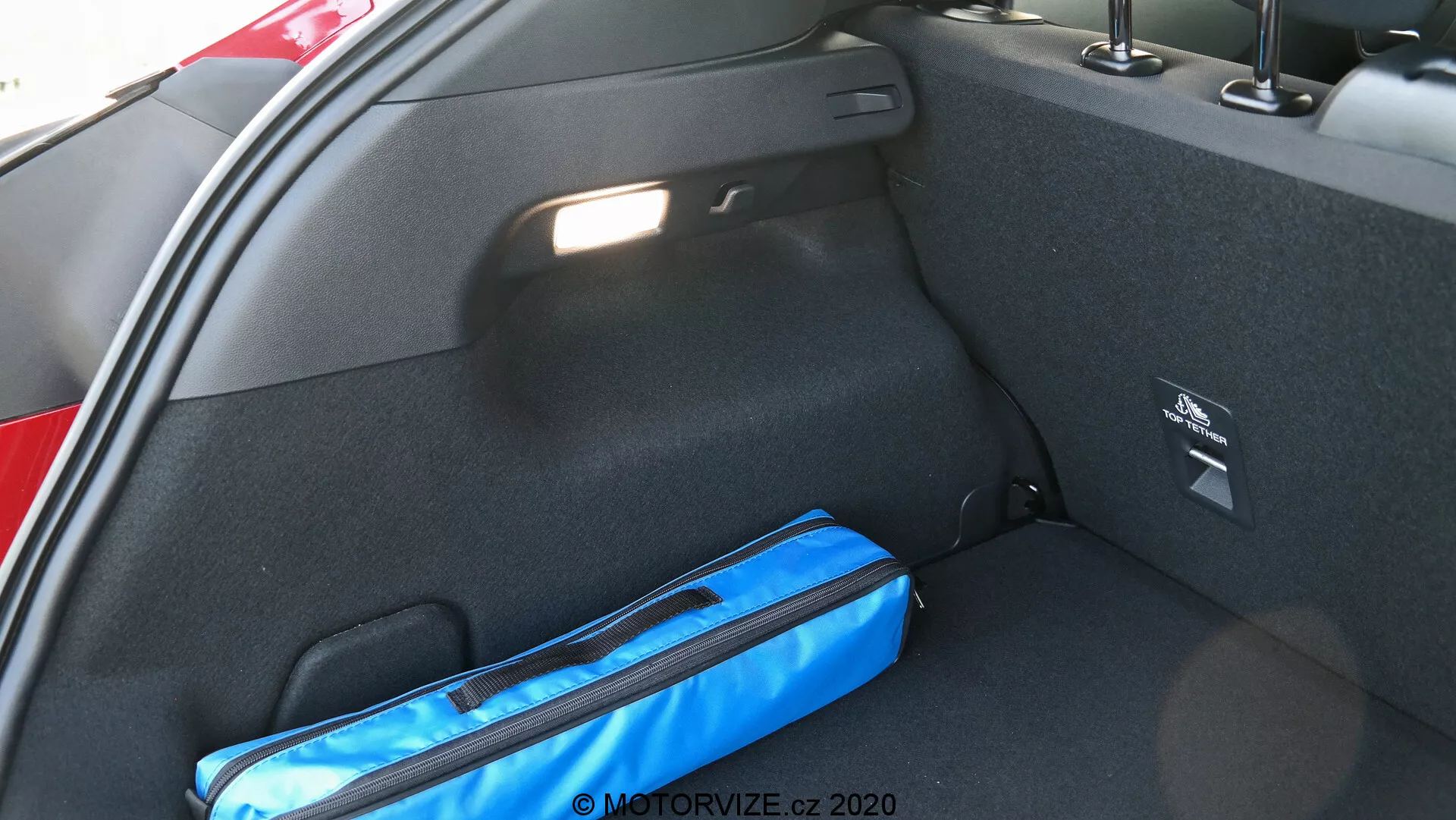 Vue rapprochée de l'intérieur du coffre d'une voiture depuis le côté, en se concentrant sur le côté droit. L'espace de chargement est en moquette noire et dispose d'un compartiment de rangement latéral avec une étiquette de sécurité. Un objet bleu, peut-être un sac ou un organiseur, repose sur le sol. La lumière naturelle éclaire le coffre, indiquant qu'il est ouvert.