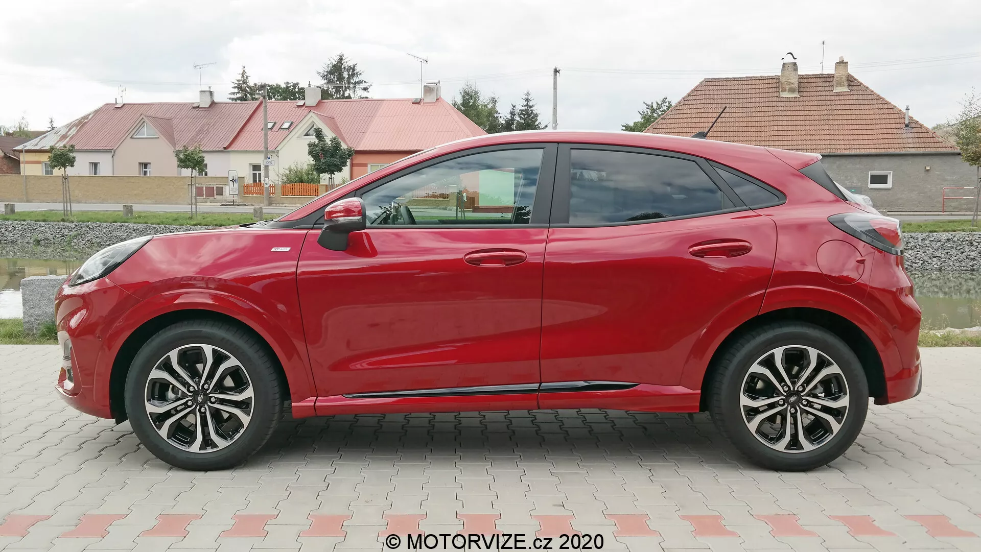 Vue de profil latéral d'une Ford Puma hatchback rouge à quatre portes 2020, prise en journée dans un parking avec en fond de l'herbe et des bâtiments résidentiels. La voiture présente des lignes de carrosserie aérodynamiques, des jantes en alliage à plusieurs rayons, des rétroviseurs et poignées de porte assortis à la couleur, et une ligne de toit descendante. L'absence de serrures visibles suggère un possible système d'entrée sans clé.