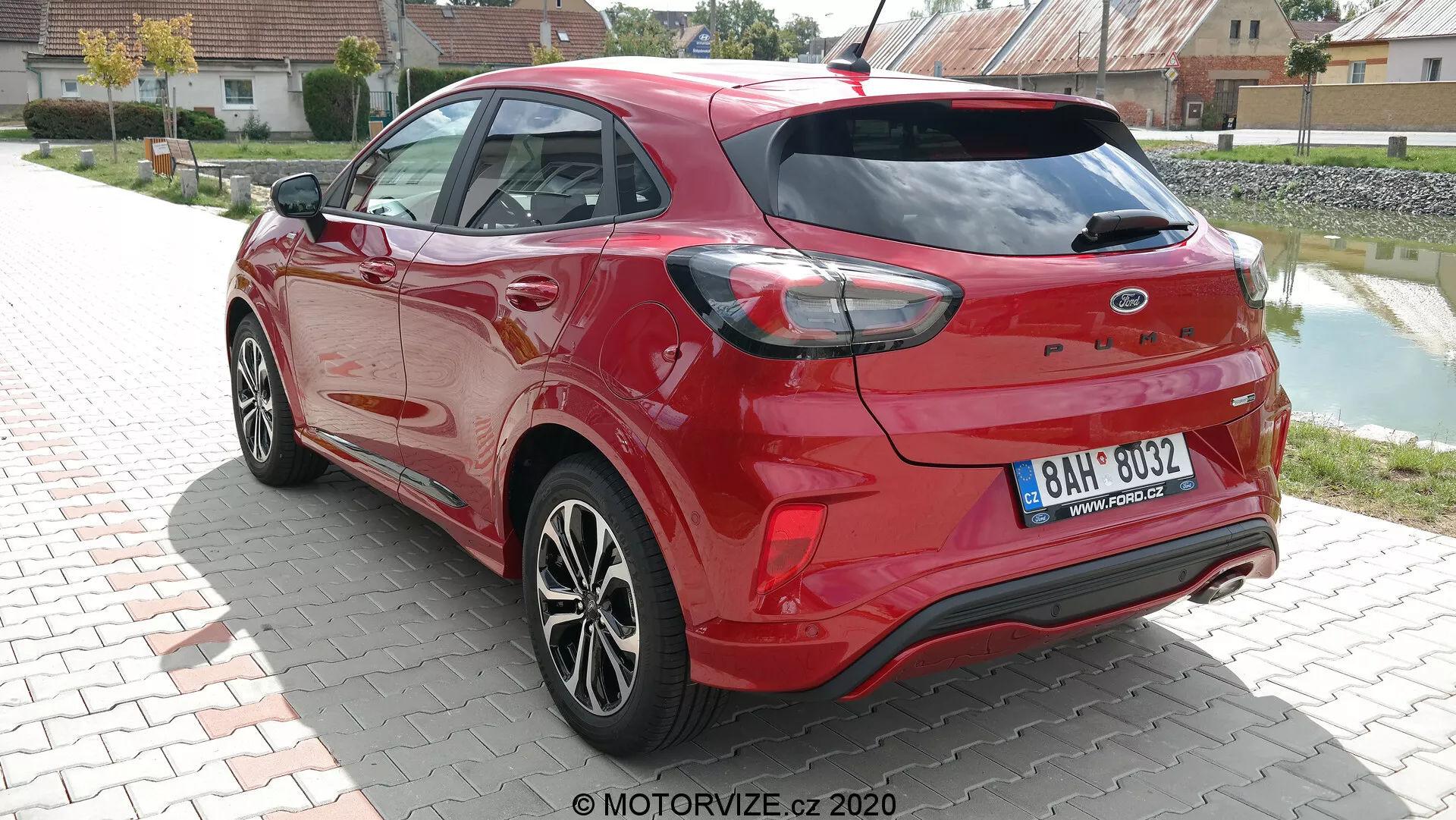 Vue arrière trois quarts d'une Ford Puma 2020 rouge vif capturée en plein jour. La prise de vue met en valeur les contours aérodynamiques de la voiture, les feux arrière distinctifs qui entourent les côtés, et les capteurs intégrés dans le pare-chocs arrière, suggérant des capacités d'assistance au stationnement. Les doubles sorties d'échappement et le spoiler arrière renforcent son attrait sportif, tandis que les jantes en alliage multi-branches et les éléments de carrosserie de la même couleur, comme les rétroviseurs latéraux et les poignées de porte, contribuent à son design moderne. Essuie-glace arrière visible, indiquant une praticité par mauvais temps.
