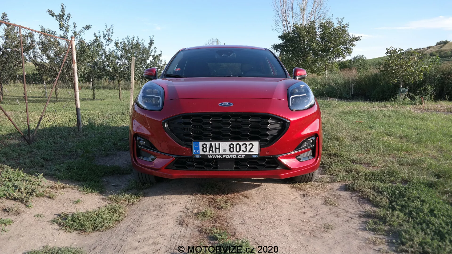Vue avant d'une Ford Puma 2020 présentant sa calandre en maille proéminente, ses phares LED élégants et les contours aérodynamiques de son capot. La voiture est positionnée sur un sol sec avec des touffes d'herbe, suggérant un cadre rural.
