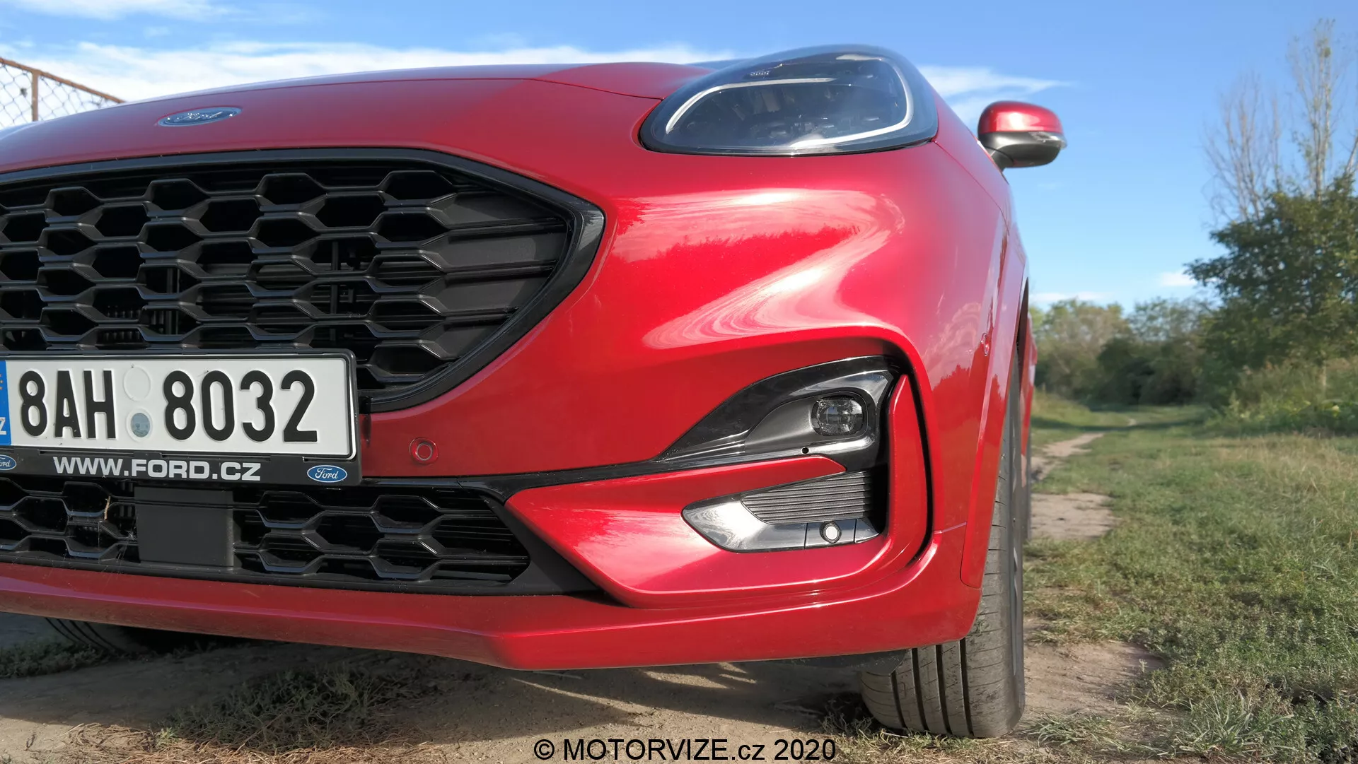 Vue en gros plan et en angle d'une Ford Puma 2020 rouge focalisée sur le quart avant gauche. Caractéristiques comprennent une grille en nid d'abeille, un phare LED/HID élégant, un pare-chocs avant et un capot aérodynamique sculpté. Une plaque d'immatriculation européenne avec une adresse web tchèque est visible. Des éléments modernes tels que les clignotants intégrés dans le rétroviseur latéral sont partiellement visibles. L'image, prise d'un angle bas, utilise la lumière naturelle et une faible profondeur de champ pour mettre en valeur le design de la voiture en extérieur.