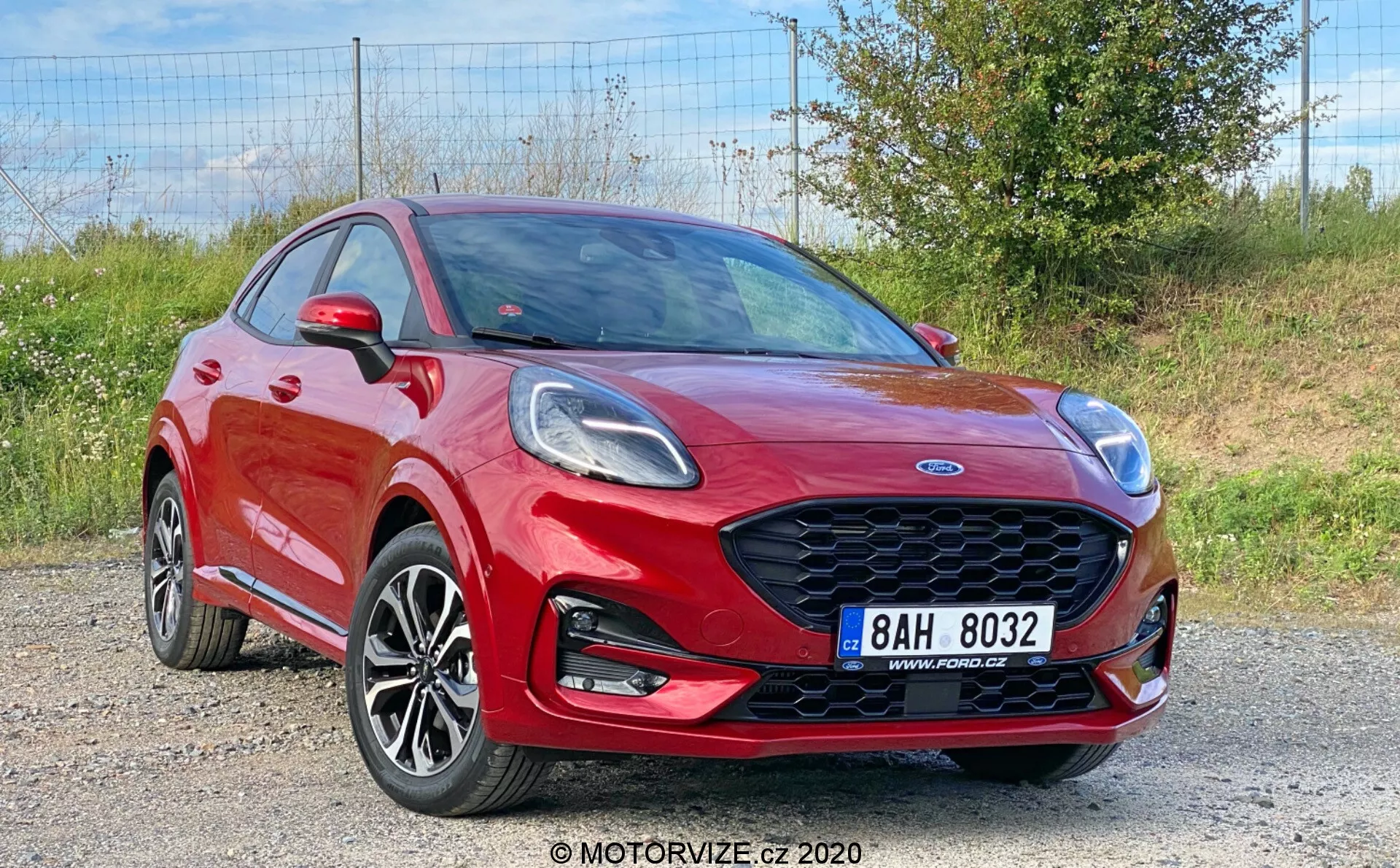L'image montre une Ford Puma 2020 rouge vif, un véhicule crossover capturé sous un angle trois quarts avant, mettant en avant sa calandre avant élégante en nid d'abeille, ses phares angulaires, ses jantes en alliage à plusieurs branches, son pare-chocs avant avec antibrouillards intégrés et sa ligne d'épaule prononcée. Le capot dynamique avec des plis, les rétroviseurs avec clignotants intégrés et une partie des portes avant et arrière sont visibles, indiquant une configuration à quatre ou cinq portes, sur un fond de gravier avec de la végétation.