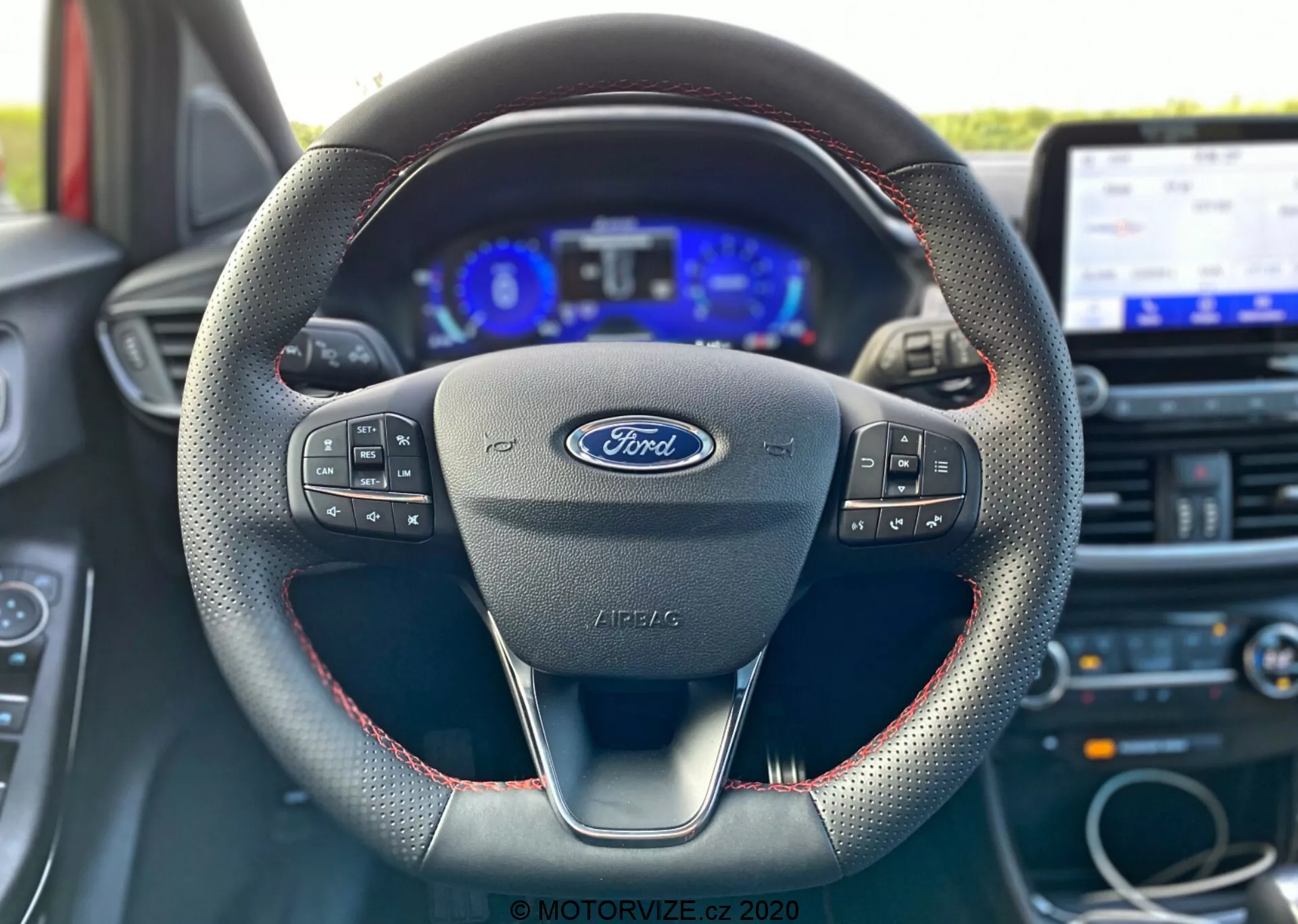 Photo intérieure d'une Ford Puma 2020 depuis la perspective du siège du conducteur, mettant en avant le volant avec logo Ford et commandes multifonctions, au design moderne à trois branches avec des coutures rouges suggérant un matériau en cuir, un tableau de bord numérique flanqué de jauges analogiques et un système d'infodivertissement à écran tactile en arrière-plan.