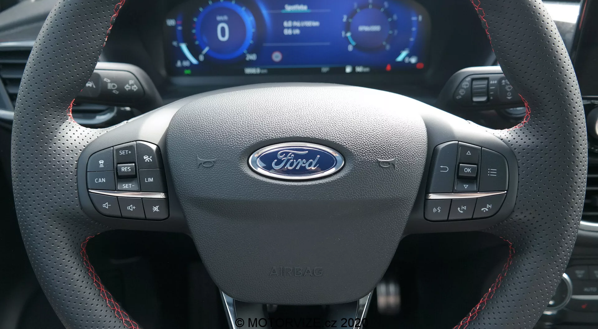 L'intérieur d'une Ford Puma 2020 vu de la perspective du conducteur montrant un volant en cuir sportif avec des coutures rouges, le logo central Ford, plusieurs boutons de contrôle, un tableau de bord numérique avec compteur de vitesse et compte-tours, et palettes de changement en métal.