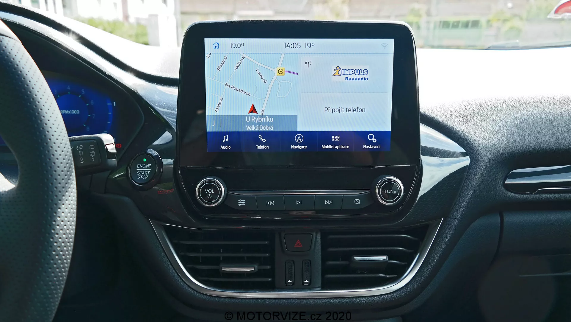 Console centrale de l'intérieur de la Ford Puma 2020 vue de la perspective du conducteur ou du passager avant, présentant le système d'infodivertissement avec écran tactile affichant la carte de navigation, boutons physiques pour les feux de détresse, les réglages de la climatisation, le lecteur CD, le port USB et la prise d'alimentation 12V. La console comporte également un thermomètre numérique affichant 17°C et une horloge affichant 14:05, le tout entouré d'éléments de design intérieur moderne avec des lignes courbes et des bouches d'aération.