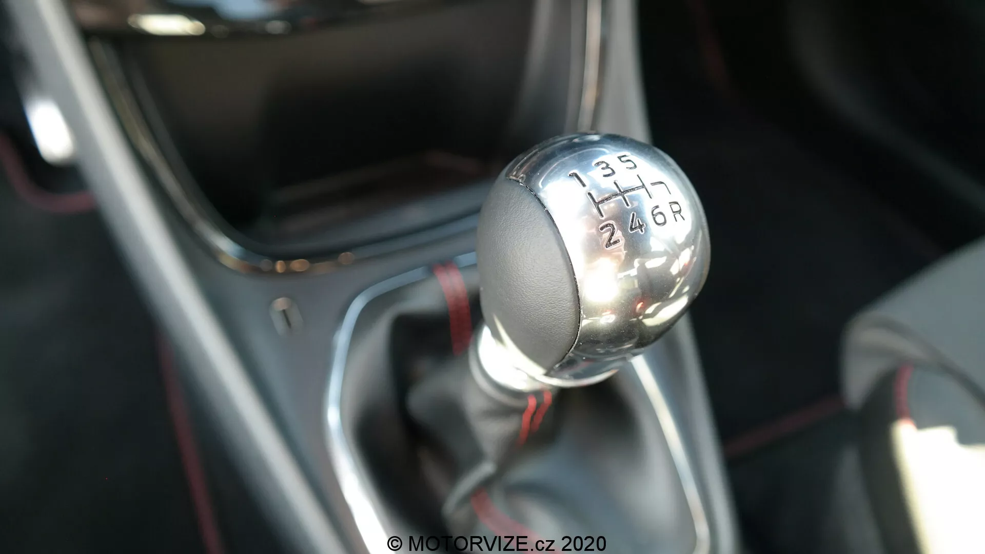 Photographie en gros plan du levier de vitesses de la transmission manuelle de la Ford Puma 2020, vue d'un angle légèrement surélevé montrant le profil supérieur et latéral. Le pommeau de vitesse métallique présente un dessus noir avec des chiffres blancs gravés indiquant un schéma de transmission à 6 vitesses avec un 'R' pour la marche arrière. Le levier de vitesses est monté sur une base avec une finition chromée et recouvert d'une gaine noire, se concentrant nettement sur le pommeau de vitesse tandis que l'arrière-plan est flou, mettant en valeur l'intérieur et la technologie de transmission du véhicule.