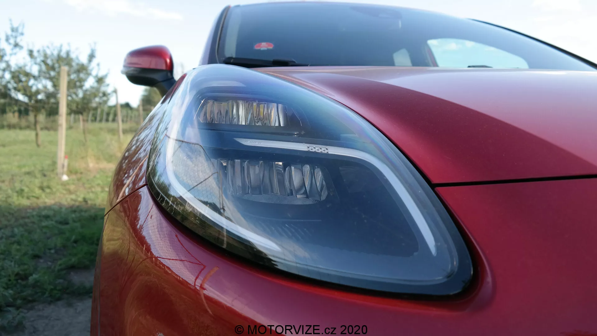Gros plan de la section avant gauche d'une Ford Puma 2020 rouge métallisé, mettant en valeur un phare moderne avec des éléments de design élégants, une partie du capot et un aperçu de la grille avant avec une faible profondeur de champ floutant l'arrière-plan, soulignant la finition extérieure premium et la technologie d'éclairage.