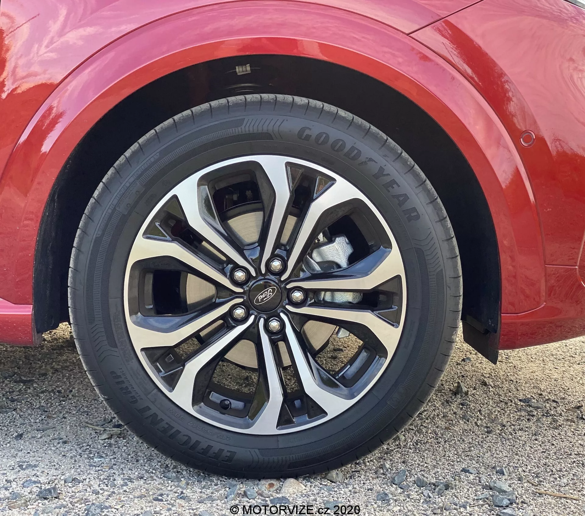 Gros plan sur une roue en alliage multi-branches sur une voiture rouge, vue sous un angle bas et légèrement incliné vers le haut. La roue présente une finition bicolore argentée et sombre, avec des branches jumelées et divisées, une surface brillante et un enjoliveur central reconnaissable. Le pneu Goodyear avec une bande de roulement substantielle et un schéma de montage à 5 écrous est également visible.