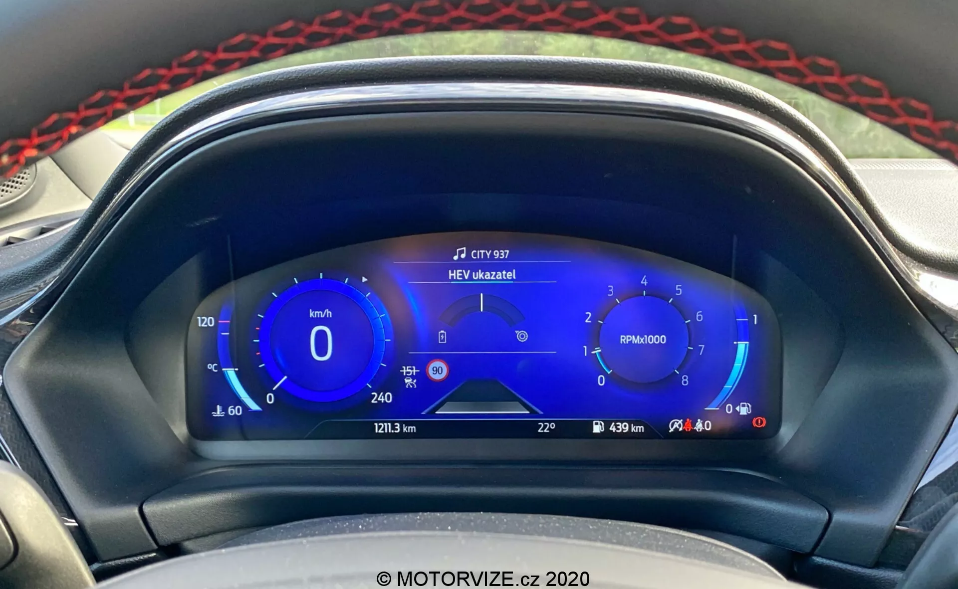 Vue rapprochée du tableau de bord numérique moderne du Ford Puma 2020 depuis le siège du conducteur. L'affichage numérique comporte un compteur de vitesse avec des plages de vitesse écologiques à gauche, un compte-tours au centre, des jauges de carburant hybrides à droite et des informations supplémentaires comme le kilométrage, la distance du trajet, l'autonomie et la température extérieure en bas. Un message en haut au centre indique 'HEV úzkazník!', signalant le mode véhicule électrique hybride. Le design élégant et axé sur la technologie, avec des nuances de bleu, blanc et rouge, met en avant les systèmes d'information avancés pour le conducteur et la technologie hybride du véhicule.