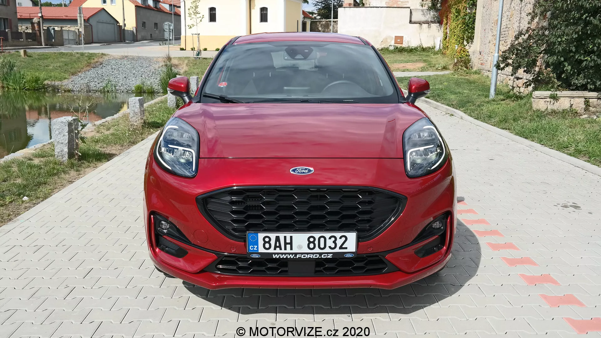 Vue de face d'une Ford Puma rouge 2020, affichant une calandre hexagonale avec grille noire, des phares effilés modernes, un pare-chocs avant sportif avec de larges prises d'air et une plaque d'immatriculation centrale. Les lignes dynamiques du capot accentuent l'esthétique du véhicule. En arrière-plan, une zone résidentielle.