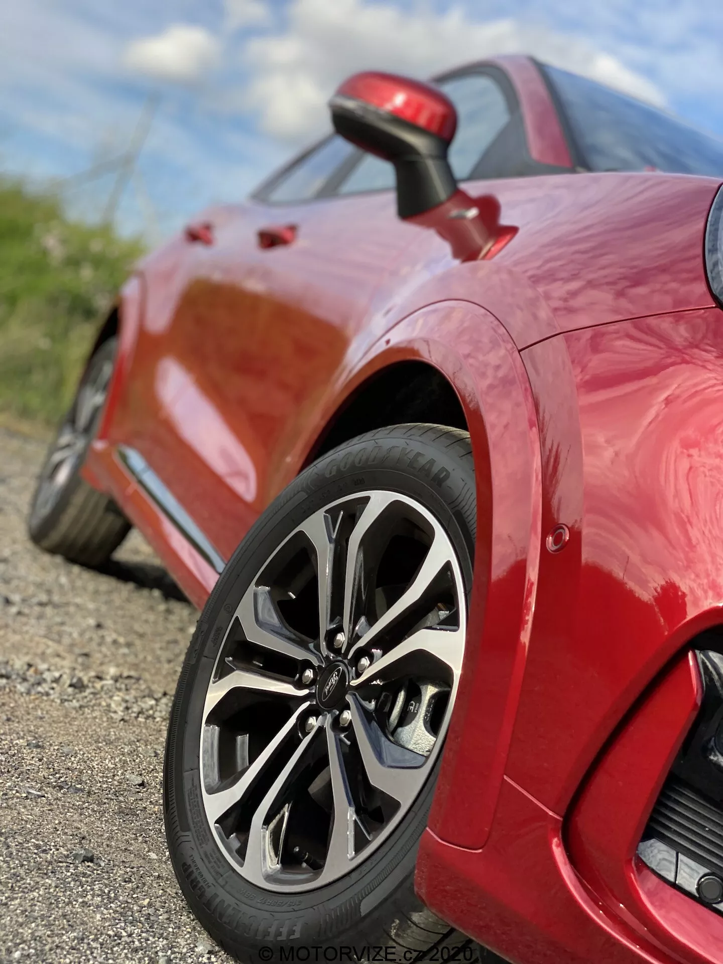 Vue rapprochée d'une voiture de sport Ford Puma 2020 rouge, centrée sur la roue arrière et le panneau latéral. La prise de vue au sol en contre-plongée met en avant le design sophistiqué de la roue à multiples rayons, le pneu à profil bas et la finition brillante de la peinture. L'image capture les détails complexes de la conception de la jante et de la bande de roulement, avec une faible profondeur de champ qui maintient la roue nette tandis que l'arrière-plan est flou. Une petite entrée d'air est visible derrière la roue sur le panneau latéral, soulignant les caractéristiques de performance et aérodynamiques du véhicule.