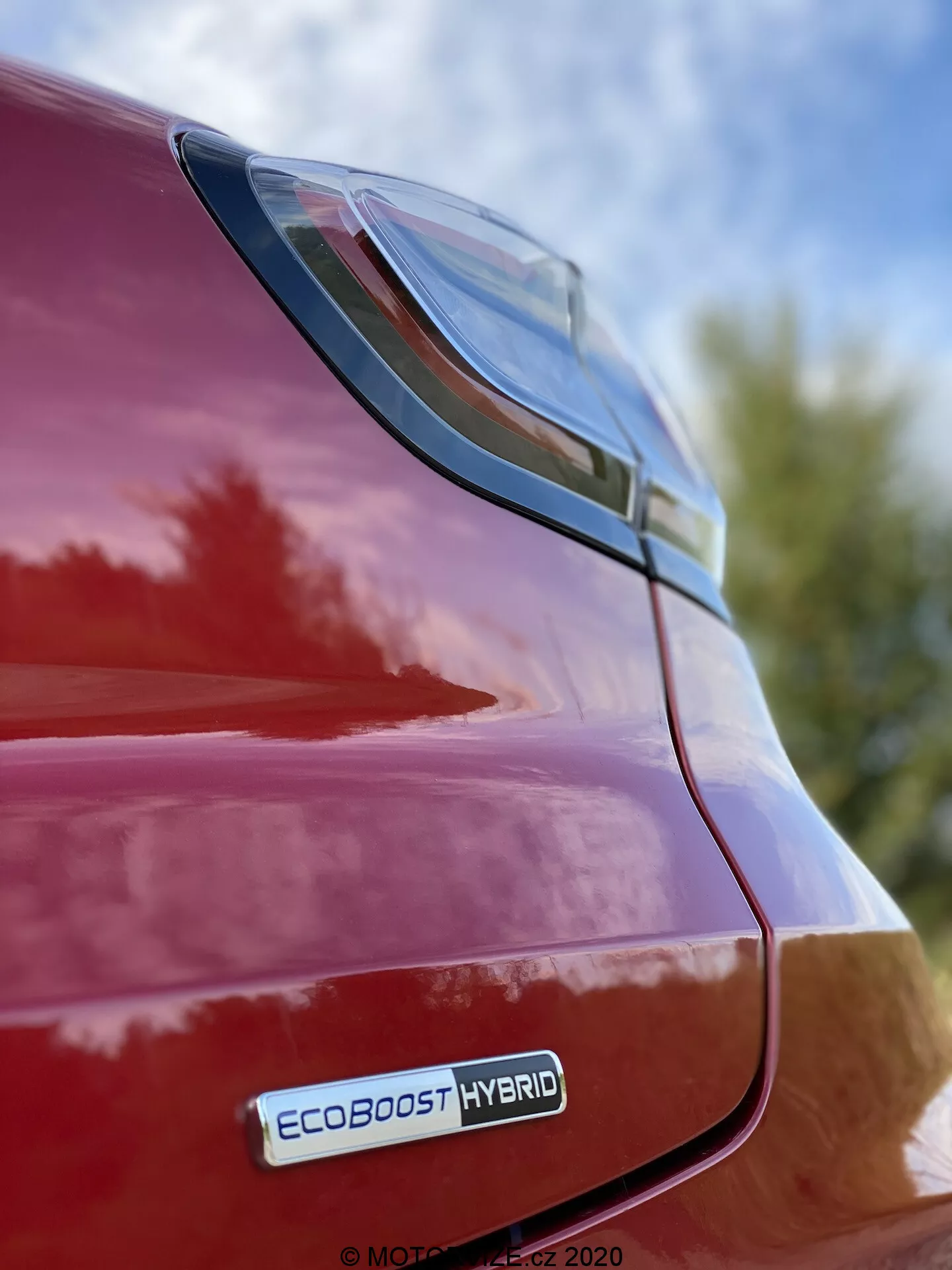 Vue rapprochée de l'arrière droit d'une Ford Puma rouge 2020, mettant en évidence le feu arrière à LED et le pare-chocs arrière depuis une perspective en contre-plongée avec l'insigne EcoBoost Hybrid visible, reflétant une finition brillante et un design élégant sur fond de ciel et de feuillage.
