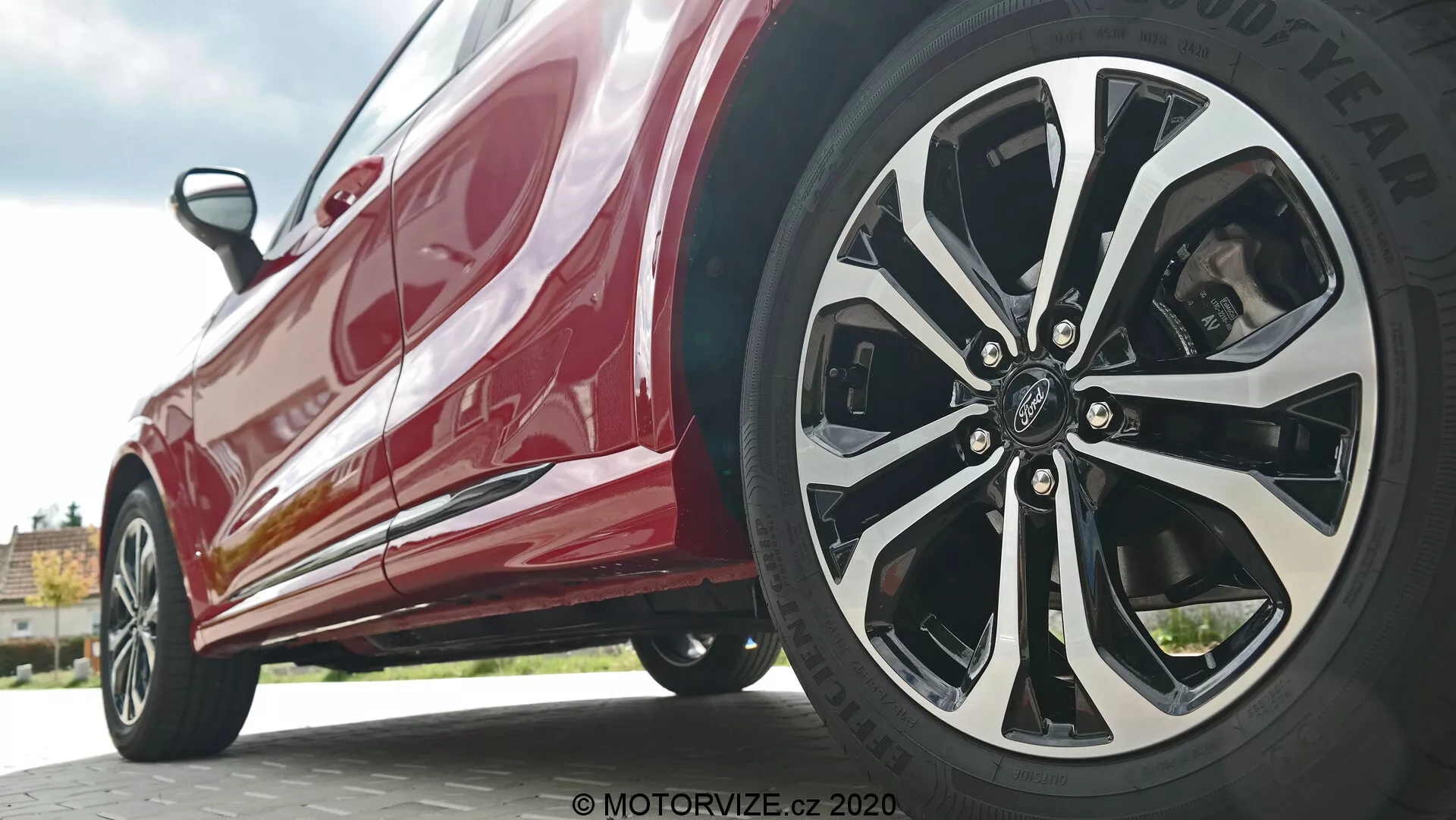 Vue rapprochée et en contre-plongée de la Ford Puma 2020 se concentrant sur le profil latéral près de la roue avant. La prise de vue depuis le niveau du sol met en évidence le design complexe à multiples rayons de la roue côté conducteur avec des finitions mélangées argent/chrome et noir, ainsi que le pneu de marque Goodyear. La carrosserie rouge et brillante reflète la lumière, montrant des éléments de design modernes tels que les jupes latérales et les lignes aérodynamiques.