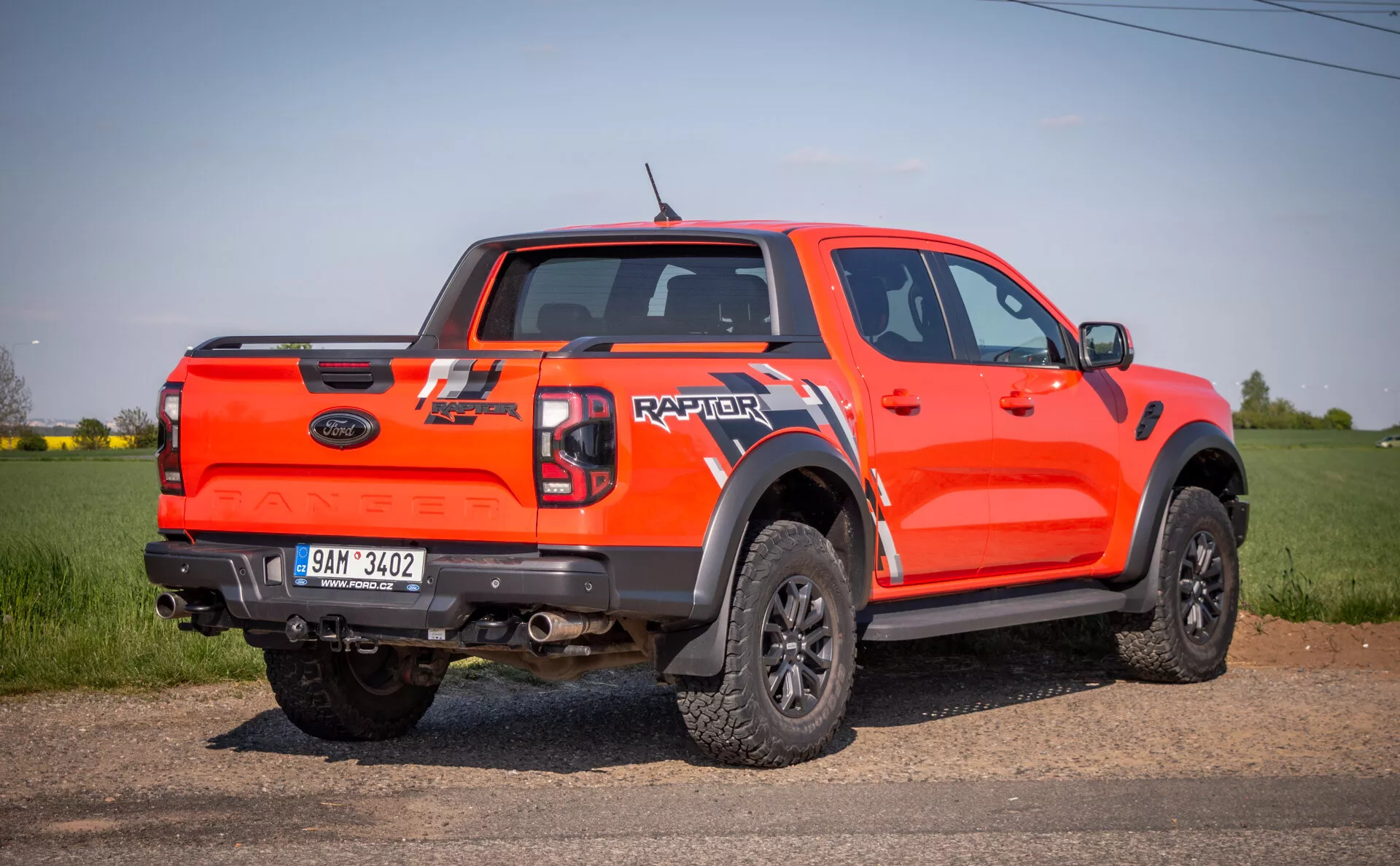 Pick-up Ford Raptor 2022 orange vif en perspective arrière-latérale, à l'extérieur sur un terrain, mettant en avant ses capacités tout-terrain avec une garde au sol élevée, cabine double, de grandes extensions d'ailes noires, jantes noires multi-branches avec pneus tout-terrain, et marquage 'RAPTOR' à l'arrière du hayon.