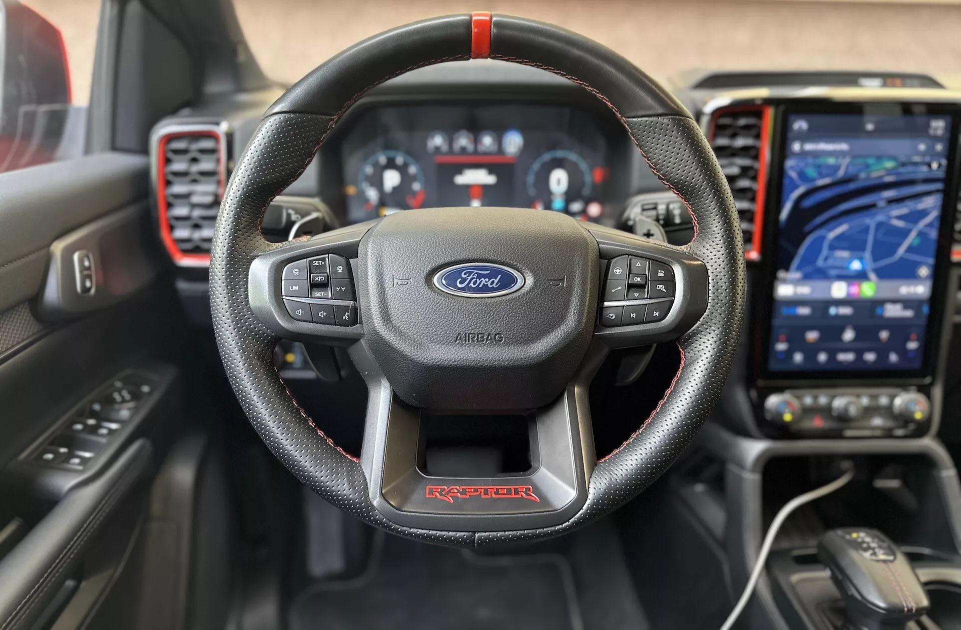 Vue avant intérieure du Ford Ranger Raptor 2022 mettant en évidence le volant en cuir à trois branches avec des coutures rouges et le logo Ford, le tableau de bord numérique, le système d'infodivertissement tactile moderne en mode navigation, le tableau de bord sportif avec des accents rouges et des aides avancées au conducteur pour une expérience de conduite de haute qualité axée sur la technologie
