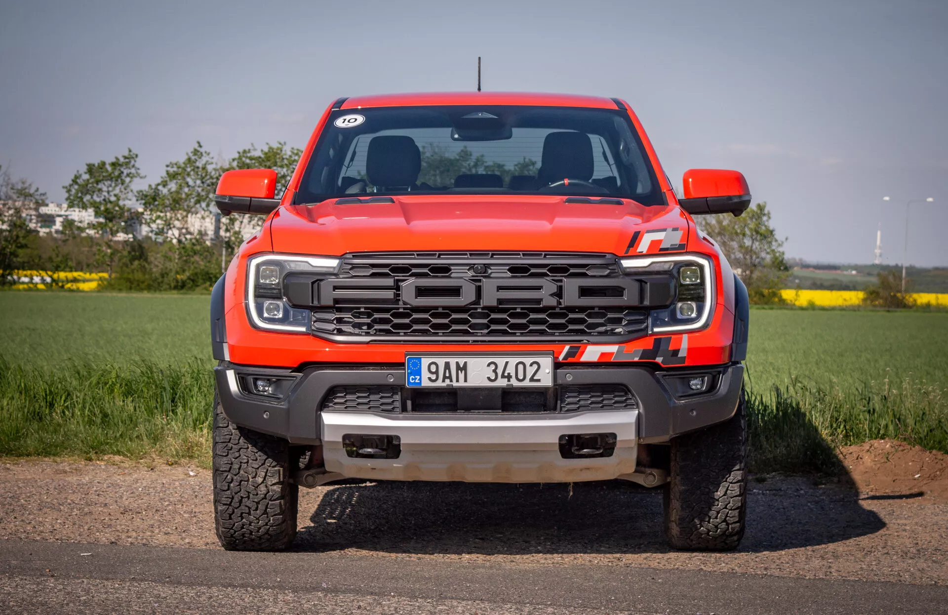 Vue de face du pickup rouge Ford Ranger 2022 sur l'asphalte avec un ciel clair en arrière-plan, mettant en avant la grille, les phares LED, le pare-chocs avant avec crochets de remorquage et plaque de protection, et le logo Ford