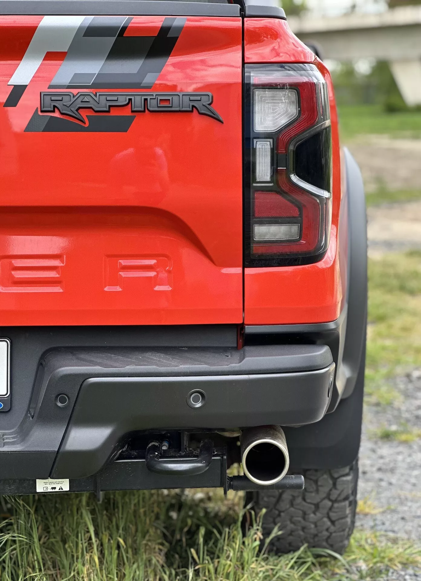 Gros plan sur le coin arrière d'un Ford Ranger Raptor rouge 2022 montrant le feu arrière à LED, le pare-chocs arrière noir avec panneau gris, l'attelage de remorque, l'embout d'échappement poli et le pneu tout-terrain, soulignant ses capacités tout-terrain et les détails de conception.