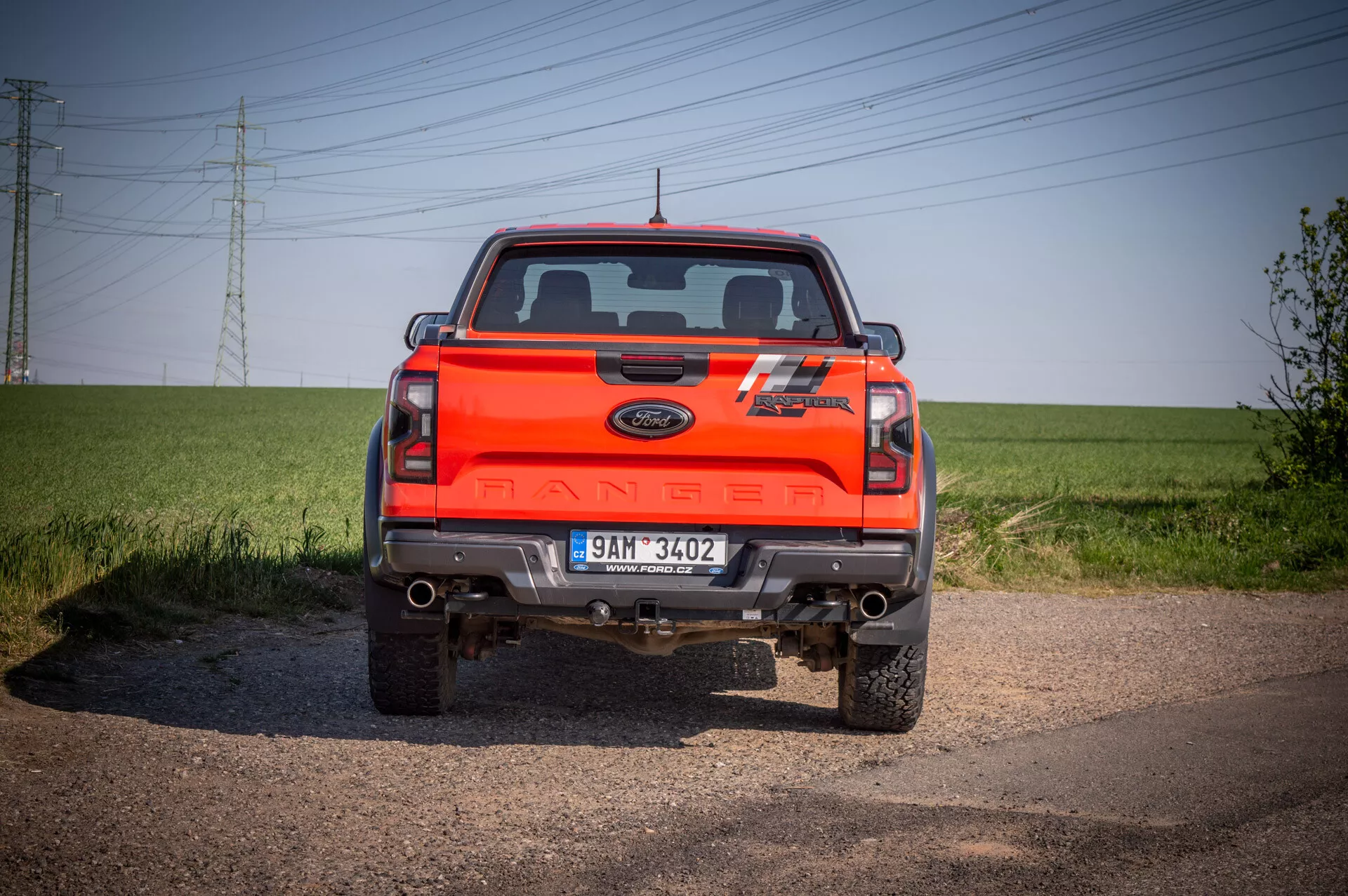 Vue arrière du Ford Ranger orange vif 2022 montrant la largeur complète, les feux arrières, le branding 'RANGER' centré et les marques d'édition spéciale, avec pare-chocs chromé présentant des capteurs de stationnement, prise sous un angle bas sur chemin de terre avec ciel clair en arrière-plan