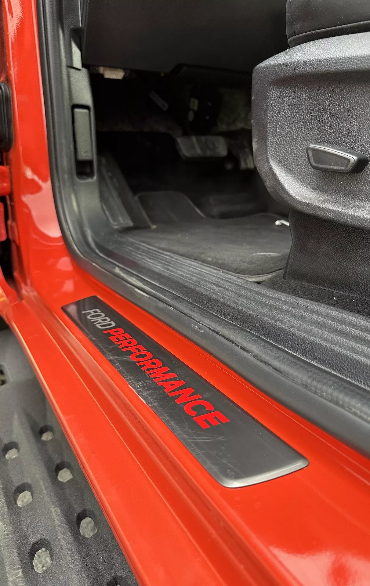 Gros plan sur le seuil de porte du Ford Ranger 2022 présentant le badge 'FORD PERFORMANCE', extérieur rouge brillant, tapis intérieur noir, siège en tissu gris au design sportif et tapis de sol en caoutchouc noir durable