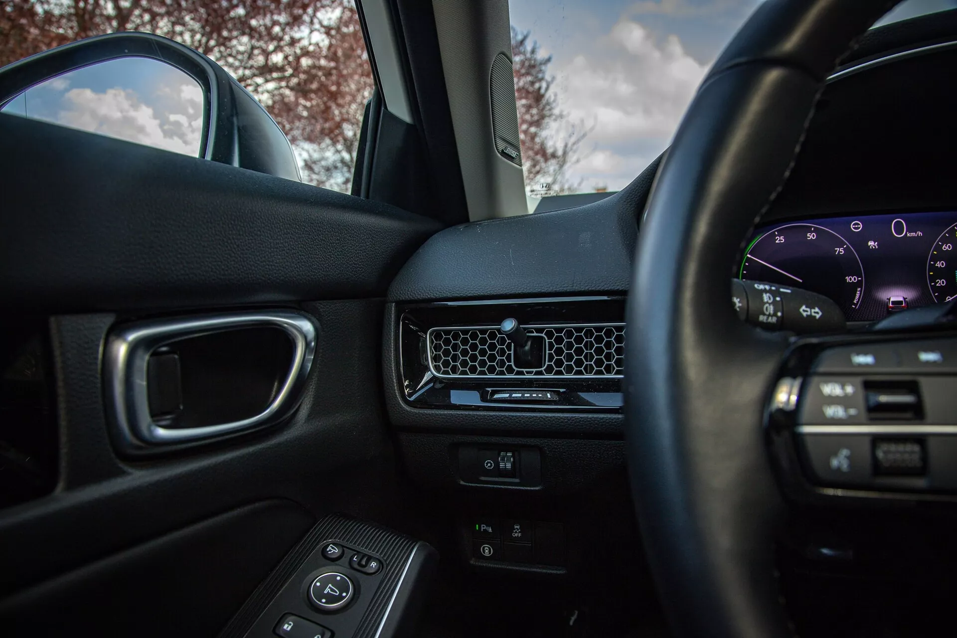 Vue intérieure du Honda Civic 2022 depuis le siège conducteur affichant le volant avec commandes multimédia, tableau de bord minimaliste avec grille d'aération en nid d'abeille, cluster d'instruments numérique, porte passager avec commandes pour les vitres électriques et siège passager avant de couleur foncée avec éclairage naturel