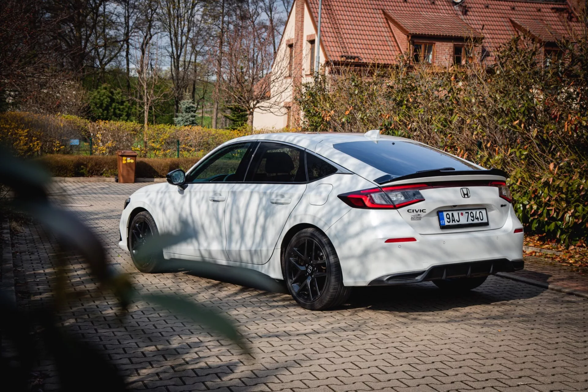 Honda Civic blanche de 2022 avec des roues multi-branches noires et un spoiler arrière, garée sur un pavé avec un feuillage automnal et une maison résidentielle en arrière-plan