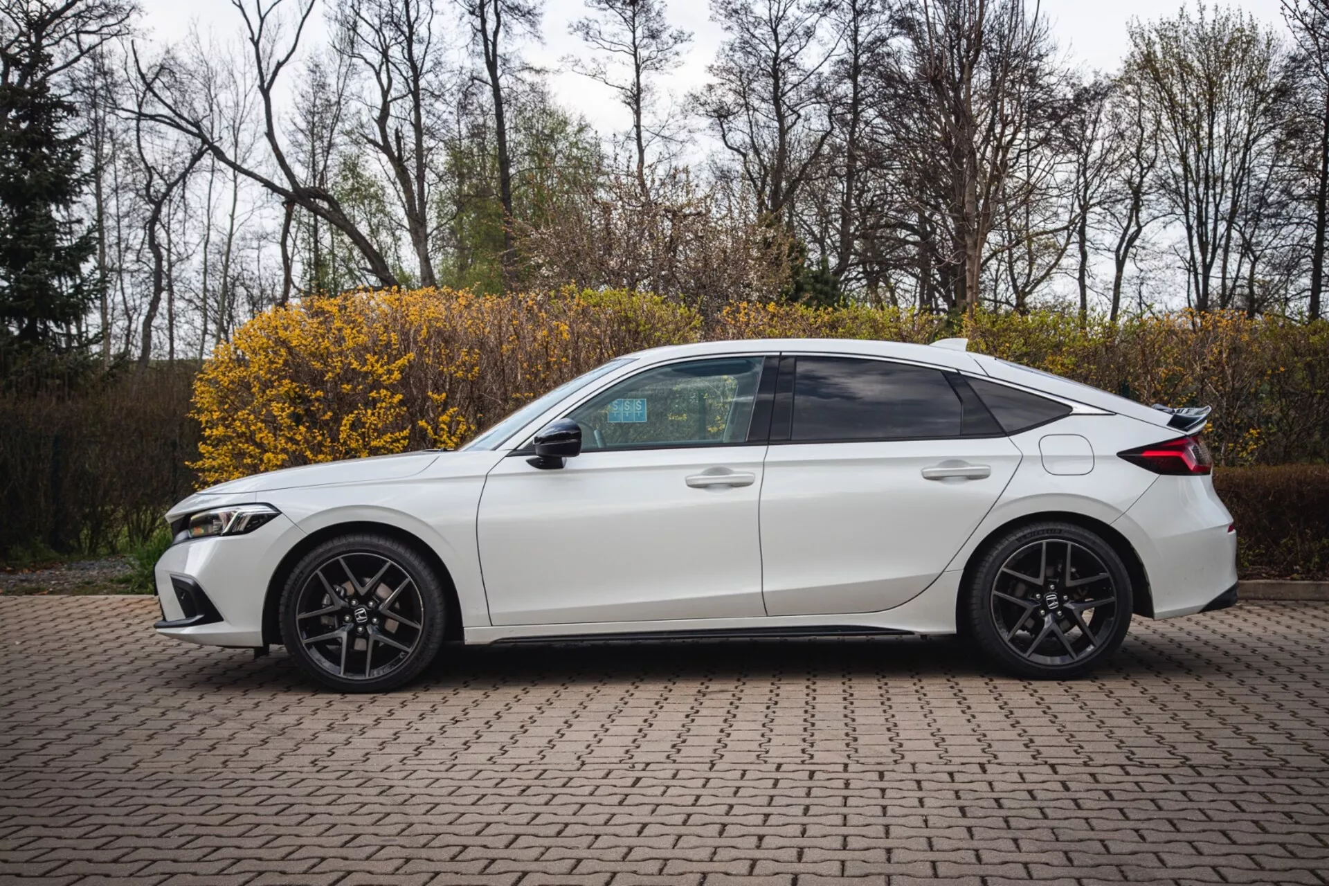 Sedan Honda Civic blanc à quatre portes de 2022 garé sur une zone pavée avec des briques imbriquées, présentant un profil latéral au design sportif avec des phares LED modernes, capturé sous un angle légèrement élevé pour mettre en évidence la vue complète du côté, y compris les fenêtres teintées, les jantes en alliage sportives et les caractéristiques technologiques telles que les clignotants intégrés dans les rétroviseurs latéraux.