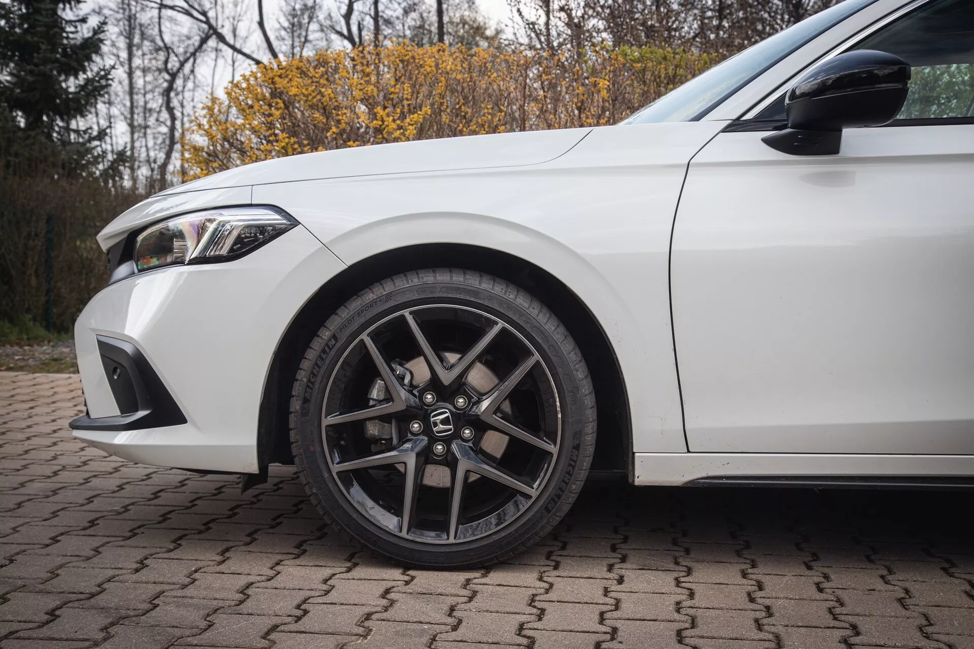 Honda Civic 2022 en blanc, montrant la roue avant gauche avec un design en alliage à plusieurs branches et pneu à profil bas, avec des détails de l'aile et une partie du pare-chocs avant sur une surface en briques