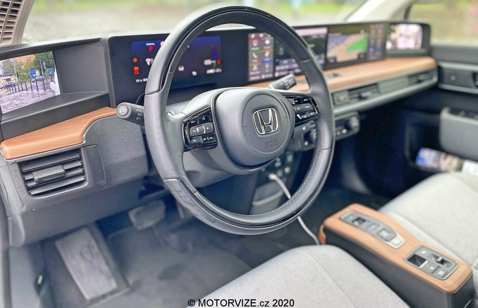 Intérieur de la Honda E 2020 présentant le côté conducteur avec un accent sur le volant et le tableau de bord moderne. Angle en gros plan mettant en évidence le logo Honda sur le volant, qui comporte des commandes pour le multimédia et le régulateur de vitesse. Les écrans allongés et continus servent de tableau de bord numérique affichant la vitesse et le système d'infodivertissement avec carte de navigation. La finition en bois visible ajoute une touche de luxe, et les sièges en tissu clair indiquent confort et style. Des fonctionnalités avancées comme les panneaux tactiles, le bouton de démarrage et le démarrage sans clé soulignent une cabine technologiquement équipée.